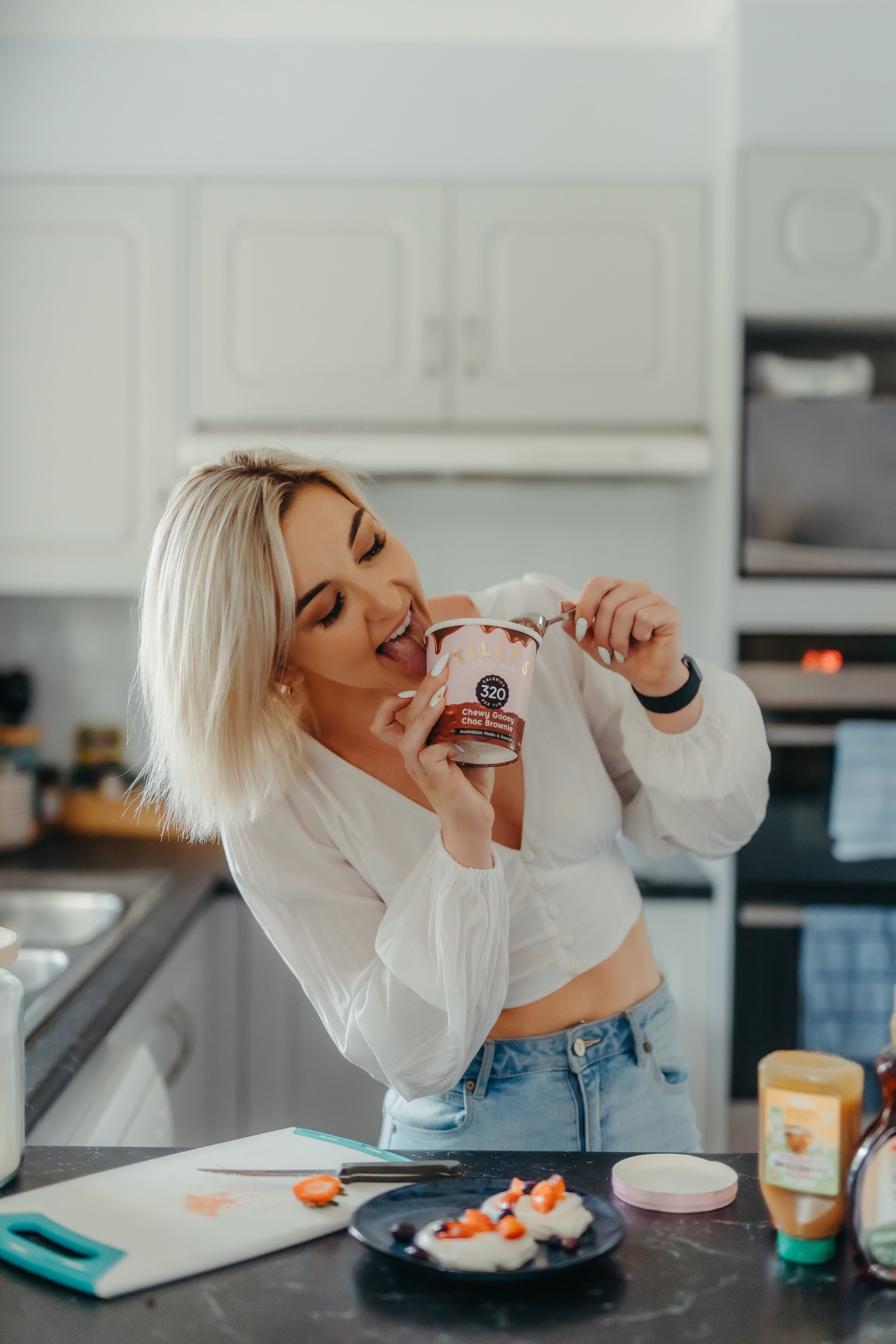 Rachel enjoying ice cream in a kitchen, wearing a white blouse and jeans, with a countertop featuring cut strawberries and kitchen items.