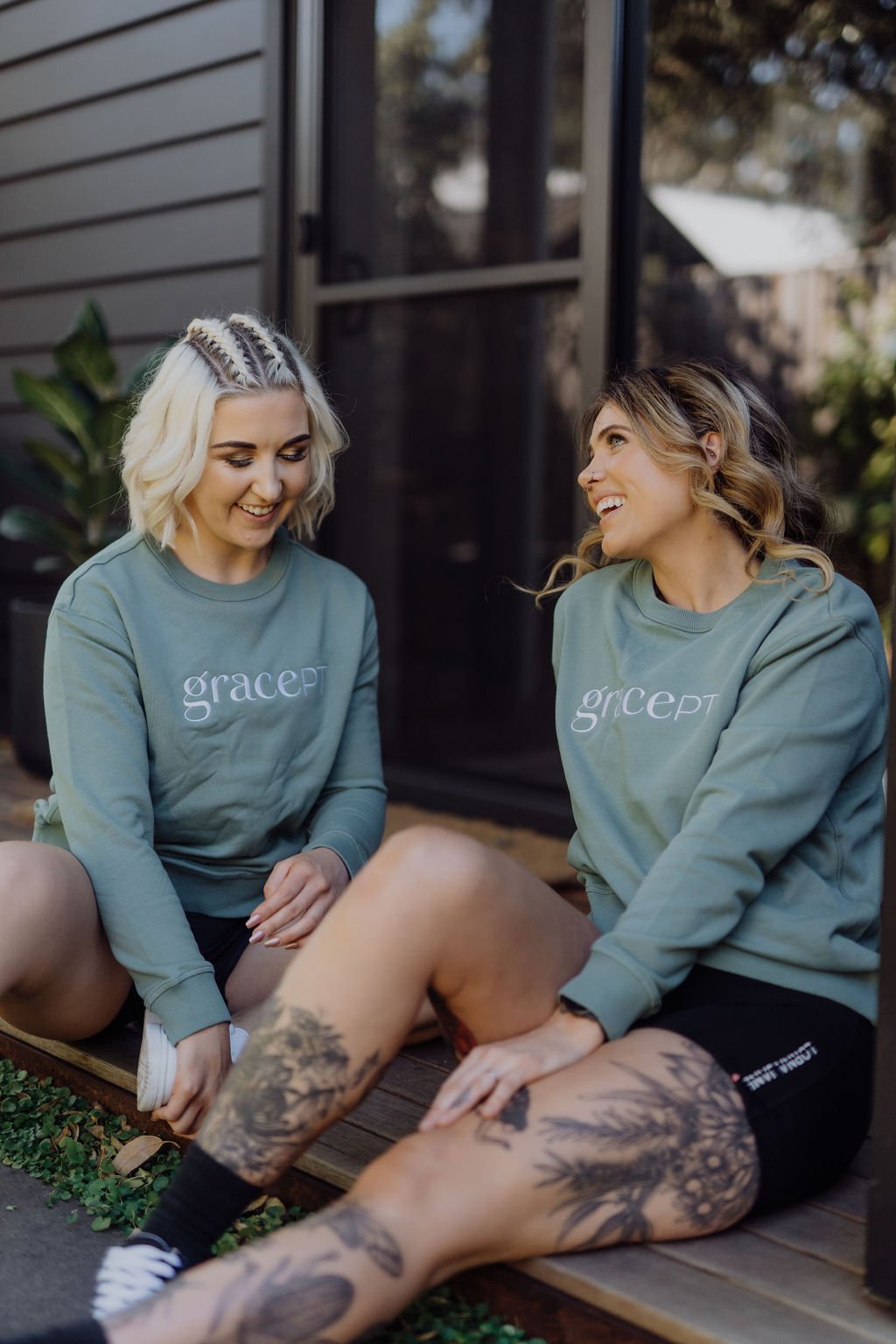 Two women sitting on steps, wearing matching green sweatshirts with the words 'grace PT.' One has tattoos on her legs, and they are both smiling and talking.
