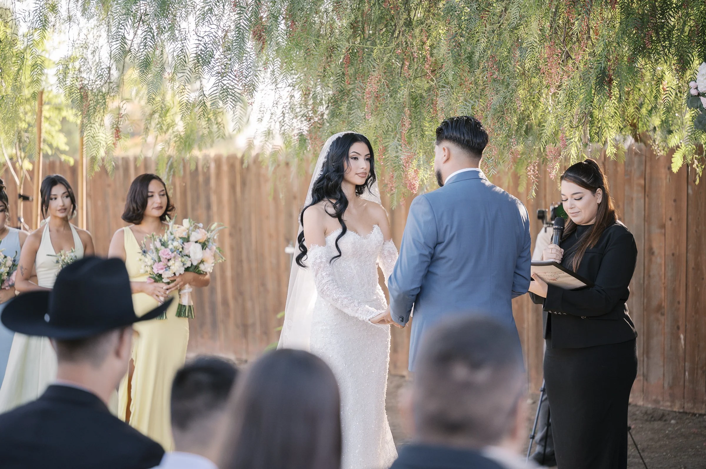 A bride in a white gown and veil holds hands with the groom in a blue suit during an outdoor ceremony. Bridesmaids in pastel dresses hold bouquets. A woman officiates under a tree, conveying a serene and joyful atmosphere.