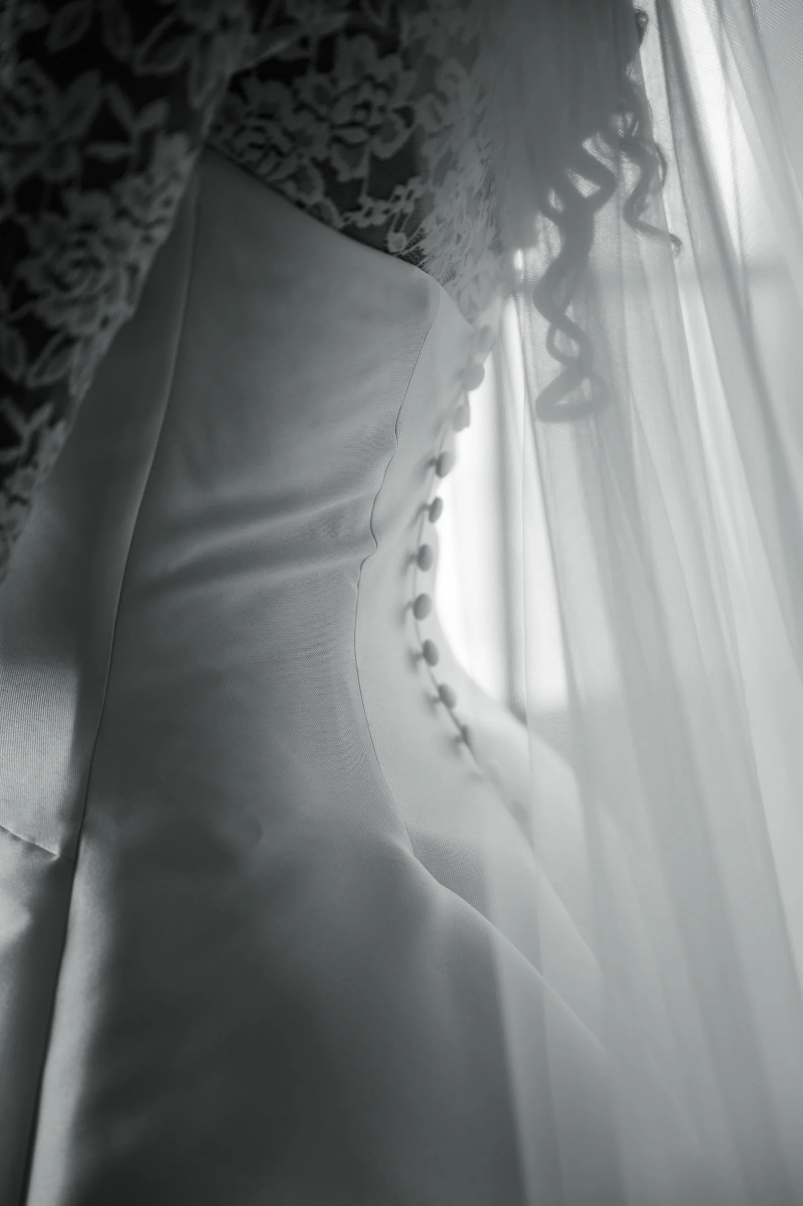 Close-up of a bride's white wedding dress in soft light, showcasing delicate lace, fabric buttons, and a veil, conveying elegance and romance.