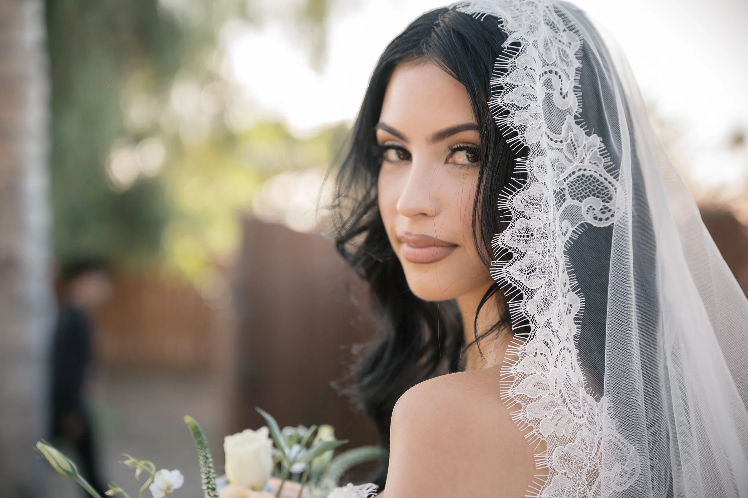 Bride in a delicate lace-edged veil smiles softly, holding a bouquet. Sunlight filters through trees, creating a warm, serene backdrop.