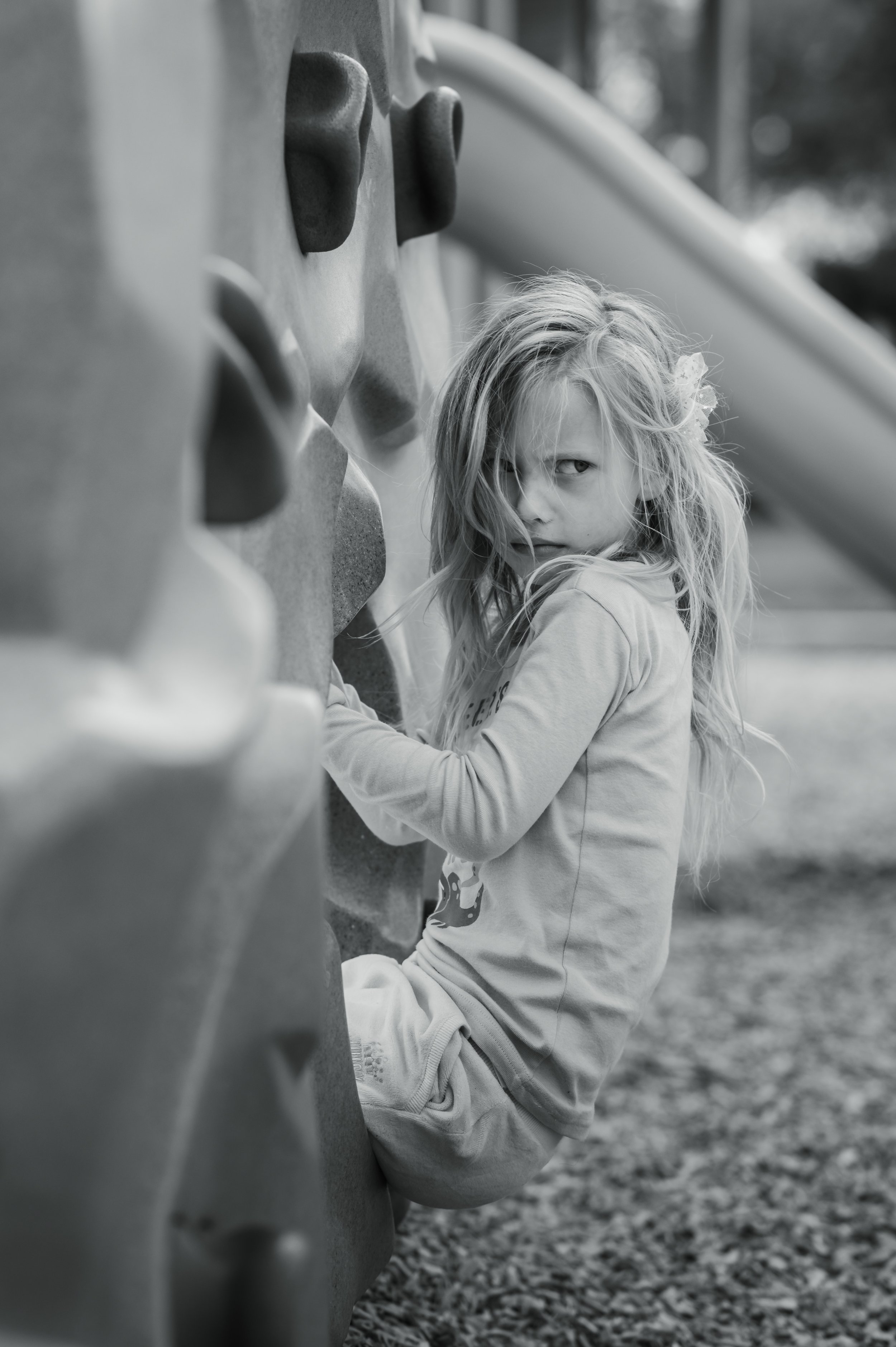 A young girl with long, tousled hair and a serious expression, sitting outdoors against a rock climbing wall, with blurred nature in the background.