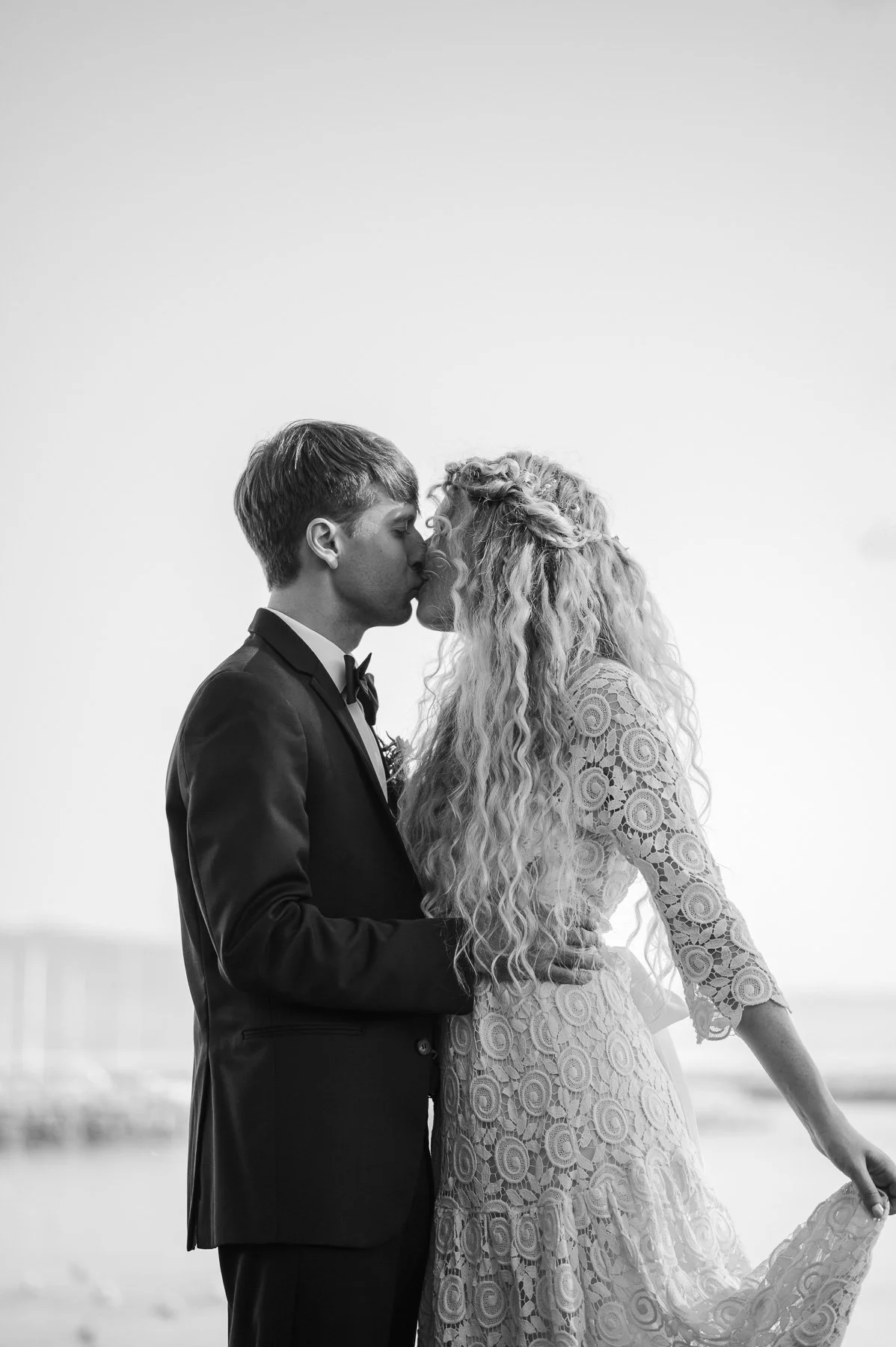 A couple kisses tenderly in black and white. The groom wears a suit, while the bride in a lace dress embraces him, capturing a romantic, timeless moment.
