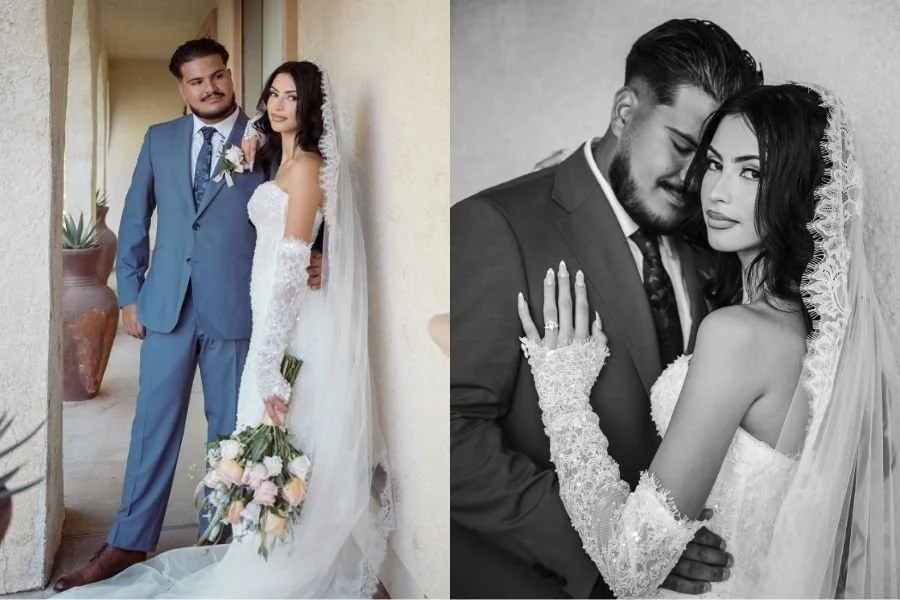A bride in a lace gown and veil stands with a groom in a blue suit, holding a bouquet of pink roses. They express joy and closeness on their wedding day.