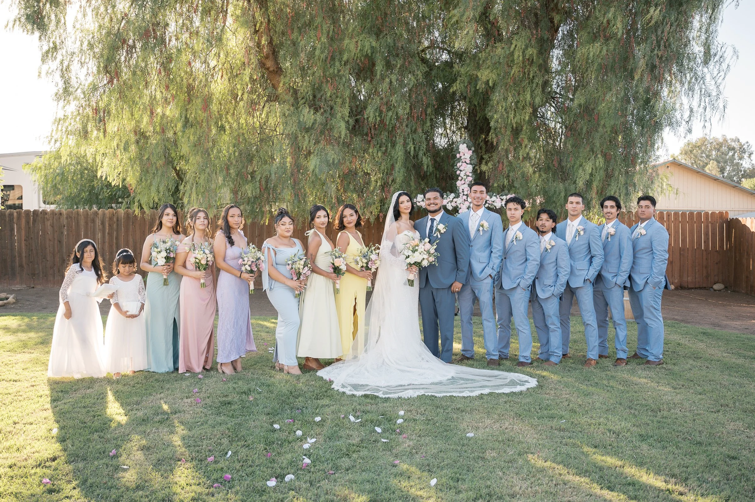 A wedding party stands on grass, with a tree behind them. The bride in a white gown and groom in a blue suit are centered, surrounded by bridesmaids in pastel dresses and groomsmen in blue suits. The mood is joyful and celebratory.