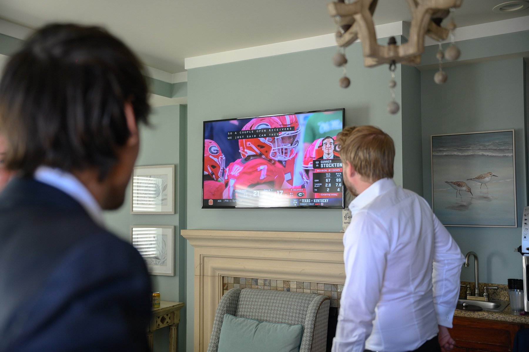 Two men in a living room watch a college football game on a wall-mounted TV. The scene feels focused and engaged, with muted decor tones.