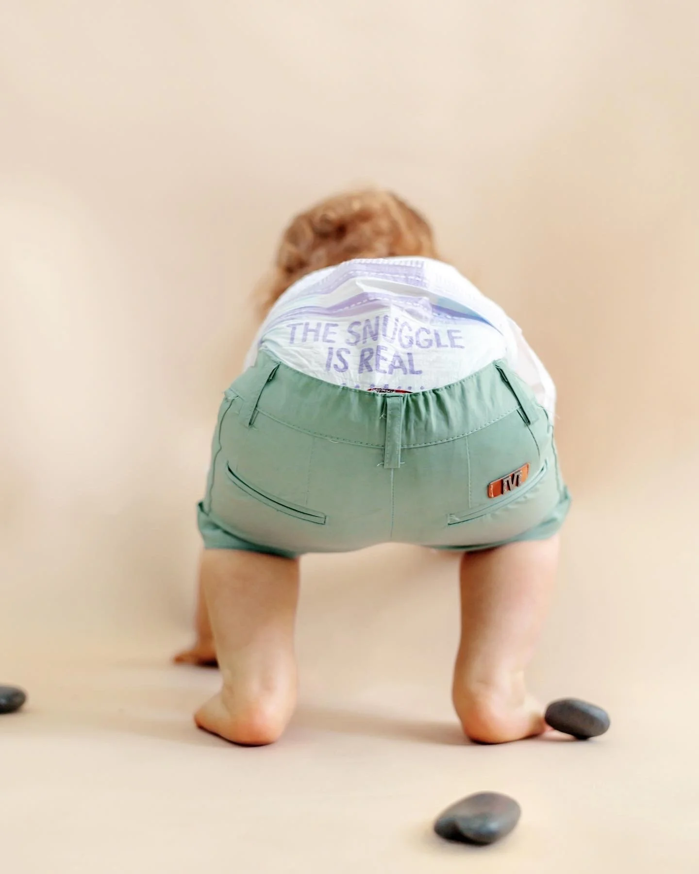Close-up of a small figurine with a person wearing green shorts and a white shirt that reads "The Snuggle is Real". The figure is crouched with hands on the ground, and rocks are placed around the feet, with a neutral background.