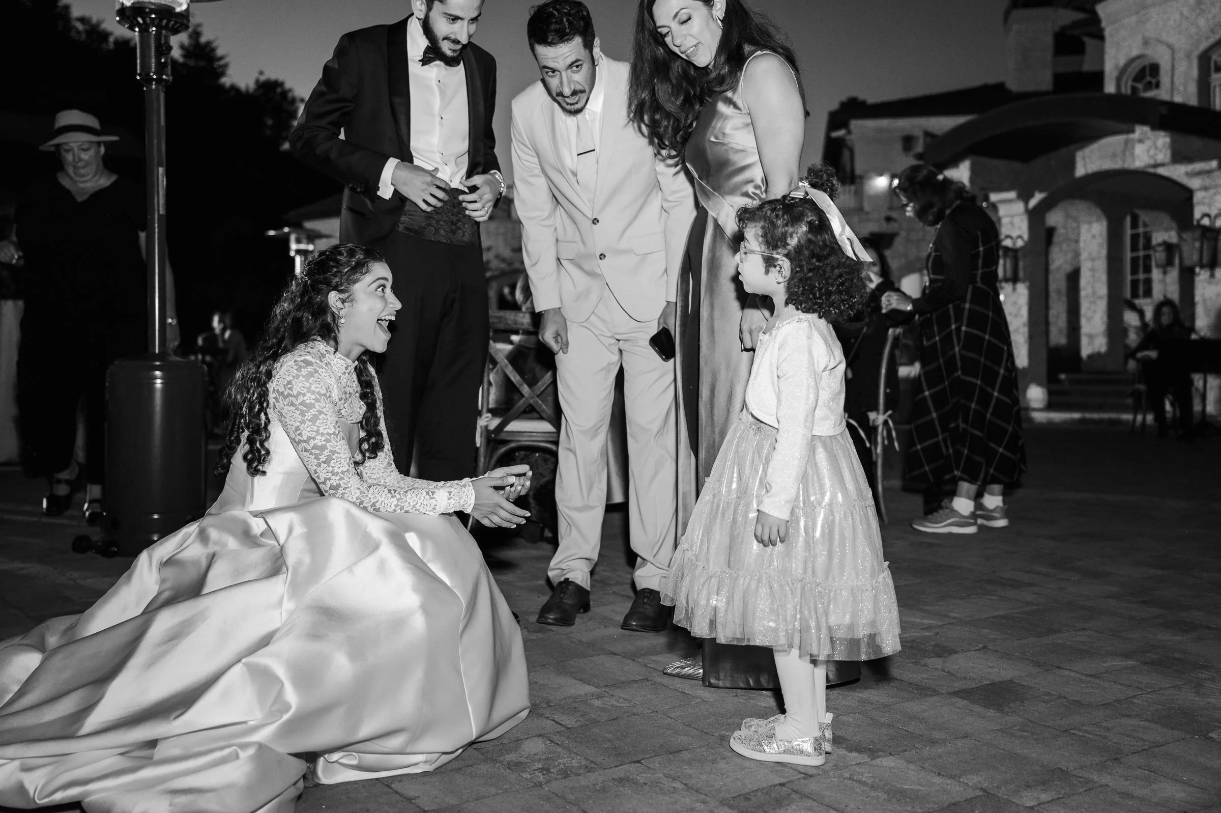 A joyful bride in a flowing gown kneels, interacting with a little girl in a sparkly dress. They are surrounded by smiling adults at an evening event.