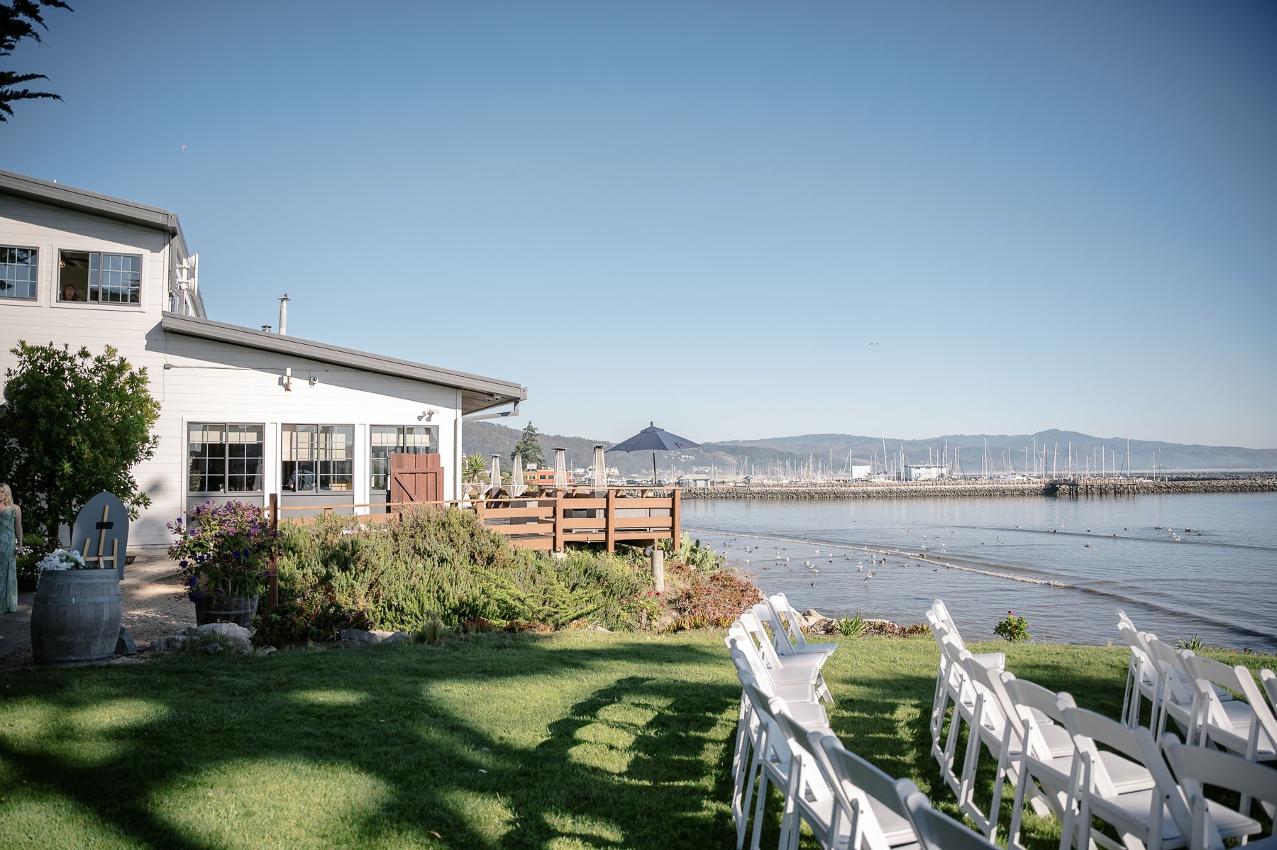 A serene waterfront venue with rows of white chairs on green grass, a wooden deck by a white building, and a calm bay under clear blue skies.