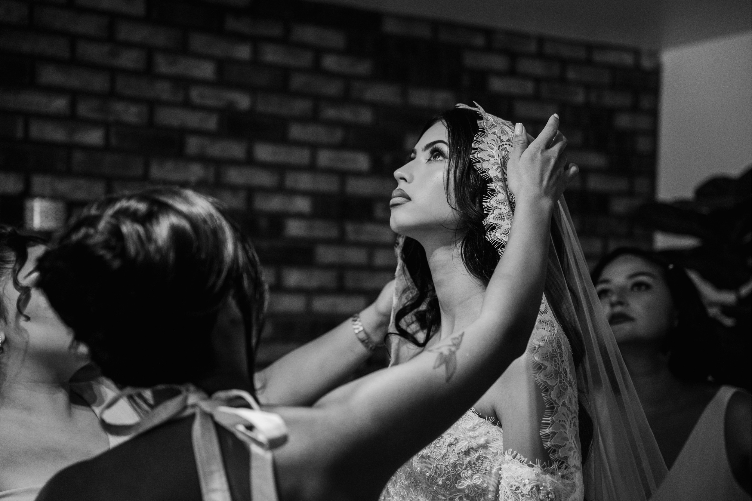 A bride gazes upwards, surrounded by bridesmaids adjusting her lace veil. The brick wall backdrop and lighting add a serene, intimate ambiance.