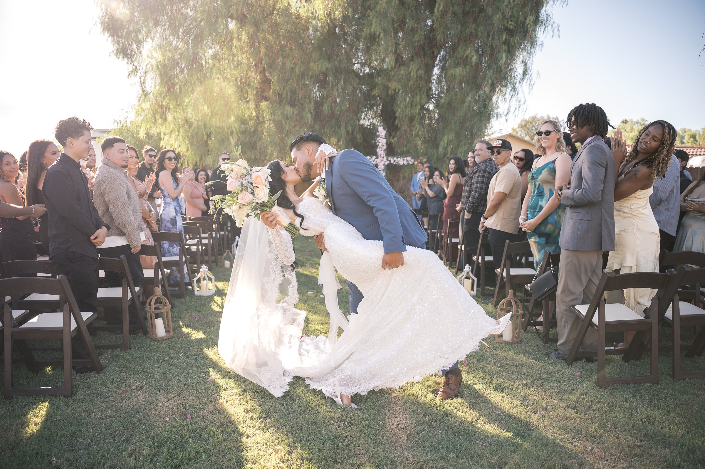 A bride and groom kiss passionately in a sunlit outdoor wedding aisle, surrounded by joyful, clapping guests. The mood is celebratory and romantic.