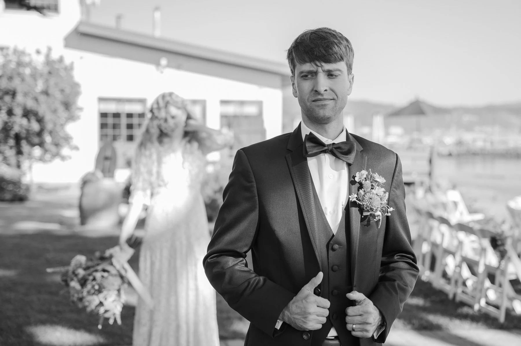 A groom in a tuxedo stands confidently outdoors, slightly smiling. Behind him, a blurred bride holds her bouquet. The mood is joyful and celebratory.