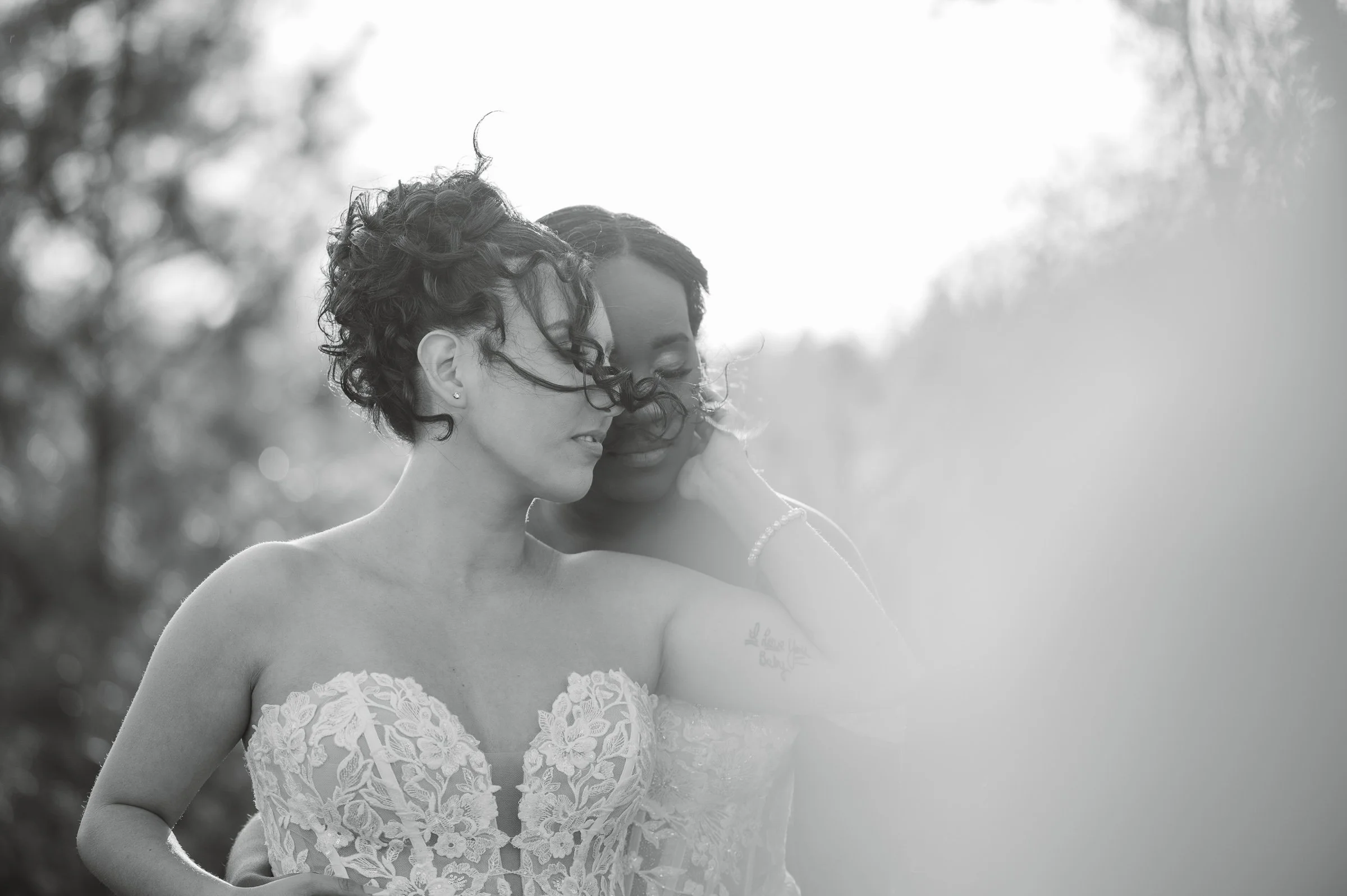 Black and white photo of two women in wedding dresses, standing close together outdoors, smiling and leaning their foreheads against each other.