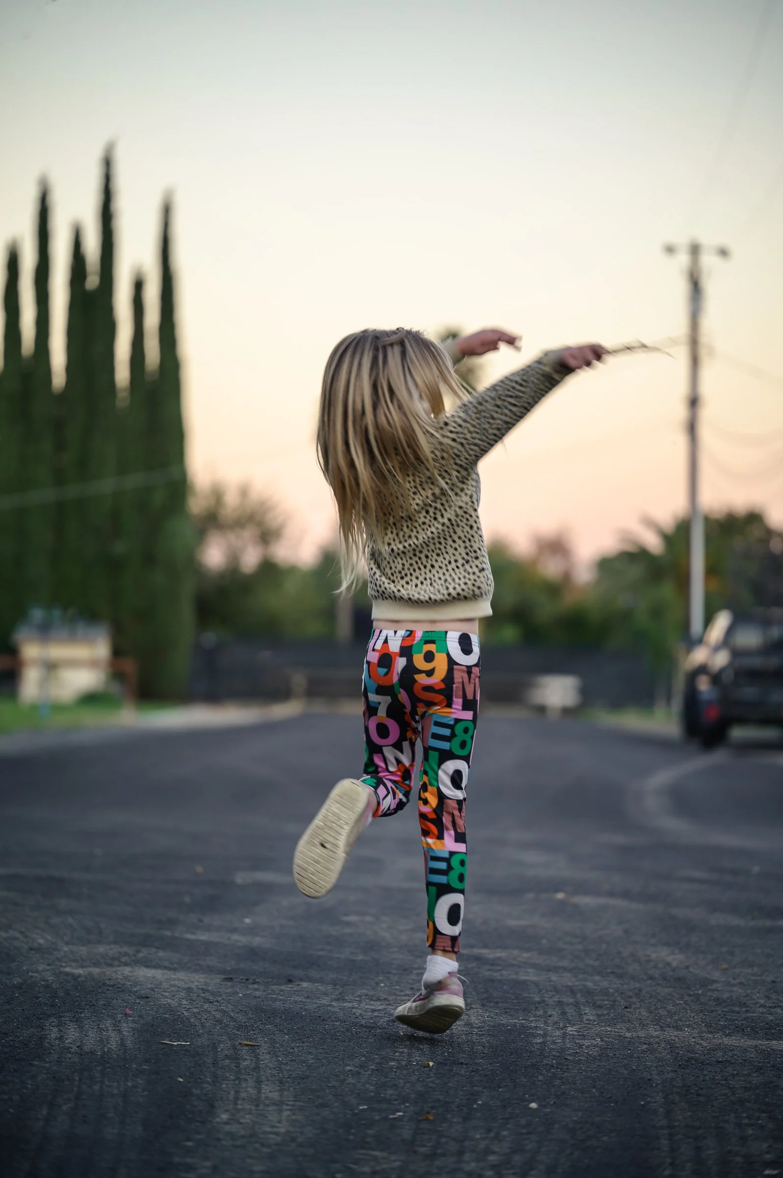 A child with blonde hair wearing colorful, lettered pants and a leopard print hoodie, running outdoors on a paved surface during sunset, with a blurry background of trees, utility pole, and parked car.