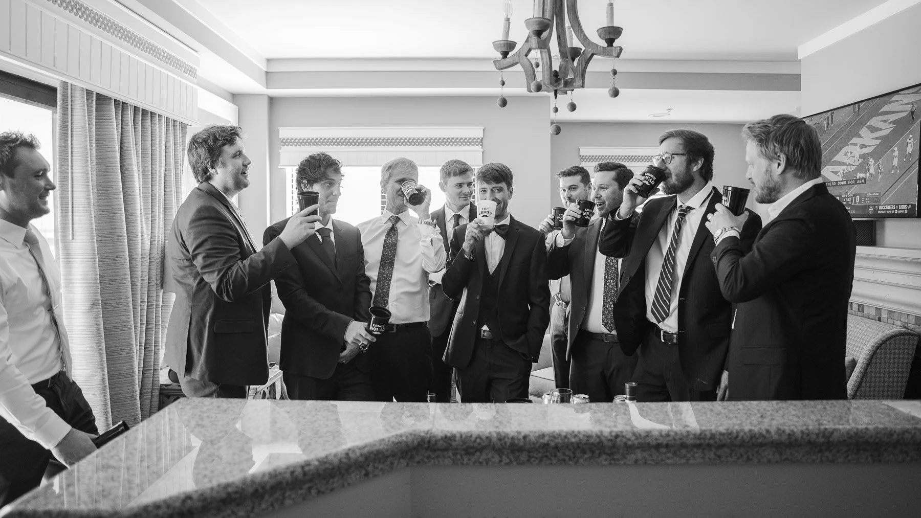 A group of men in suits stand together in a room with a chandelier, holding drinks and smiling, conveying a celebratory and jovial atmosphere.