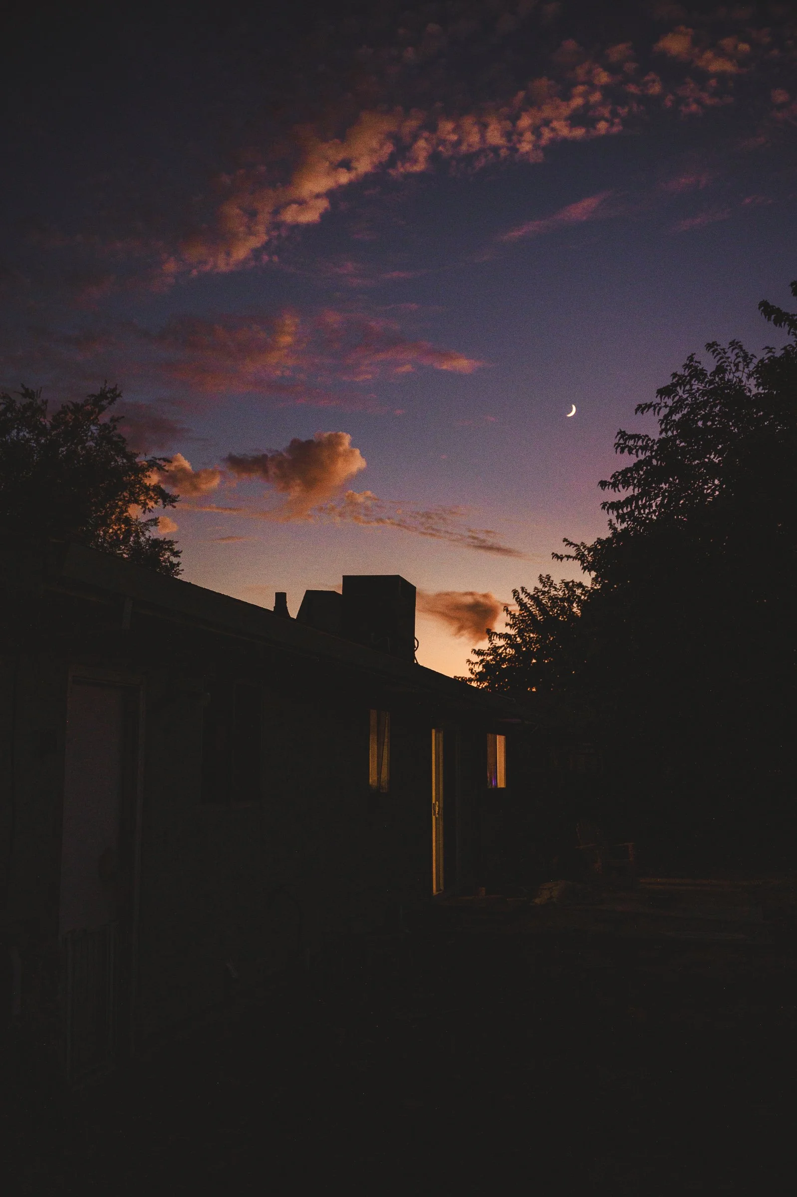 Sunset sky with a crescent moon, clouds illuminated with pink and orange hues, silhouettes of trees, rooftops, and chimney pipes in the foreground.
