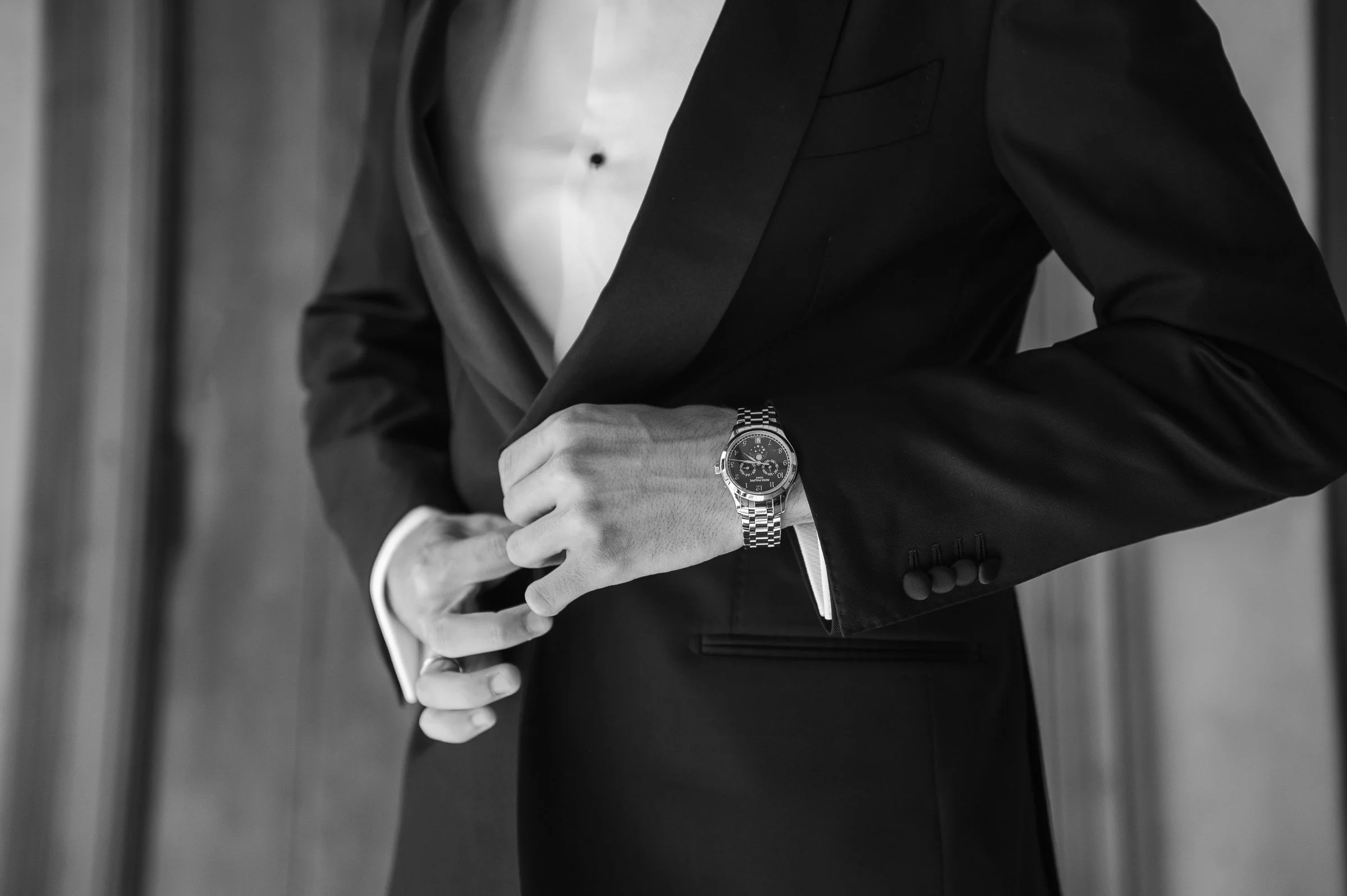 Man wearing a tailored black tuxedo, adjusting his cuff with a silver watch visible. The image conveys elegance and sophistication. Black and white tone.
