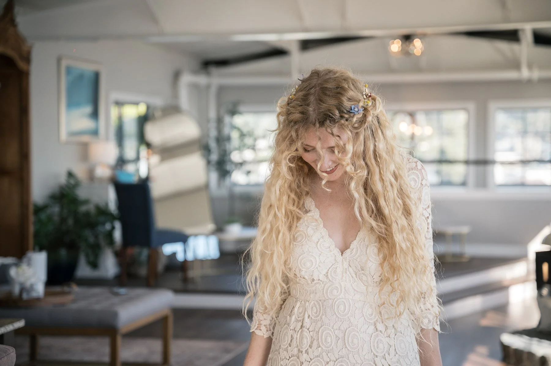A woman with long curly blonde hair adorned with small flowers looks down, smiling softly, wearing a lace dress in a bright, cozy room with soft lighting.