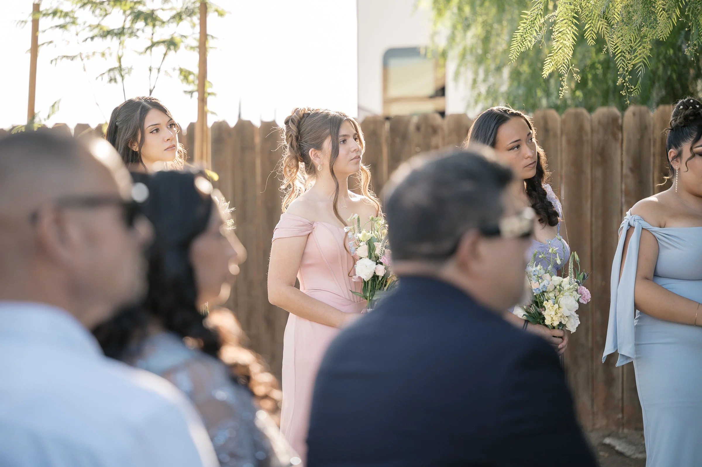 A group of bridesmaids in pastel dresses, holding bouquets, watch a ceremony attentively outdoors. Backlit, with soft sunlight and a wooden fence backdrop.