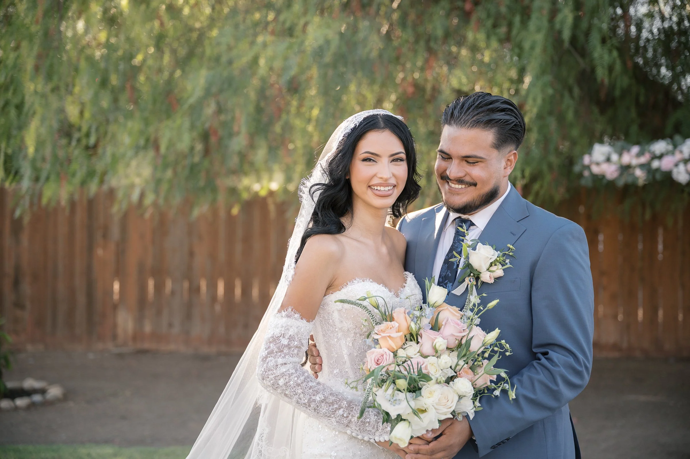 A joyful couple on their wedding day stands outdoors. The bride, in a lace gown, holds a bouquet of pink and white flowers. The groom in a blue suit smiles warmly. A wooden fence and greenery form the backdrop, conveying a serene, happy atmosphere.