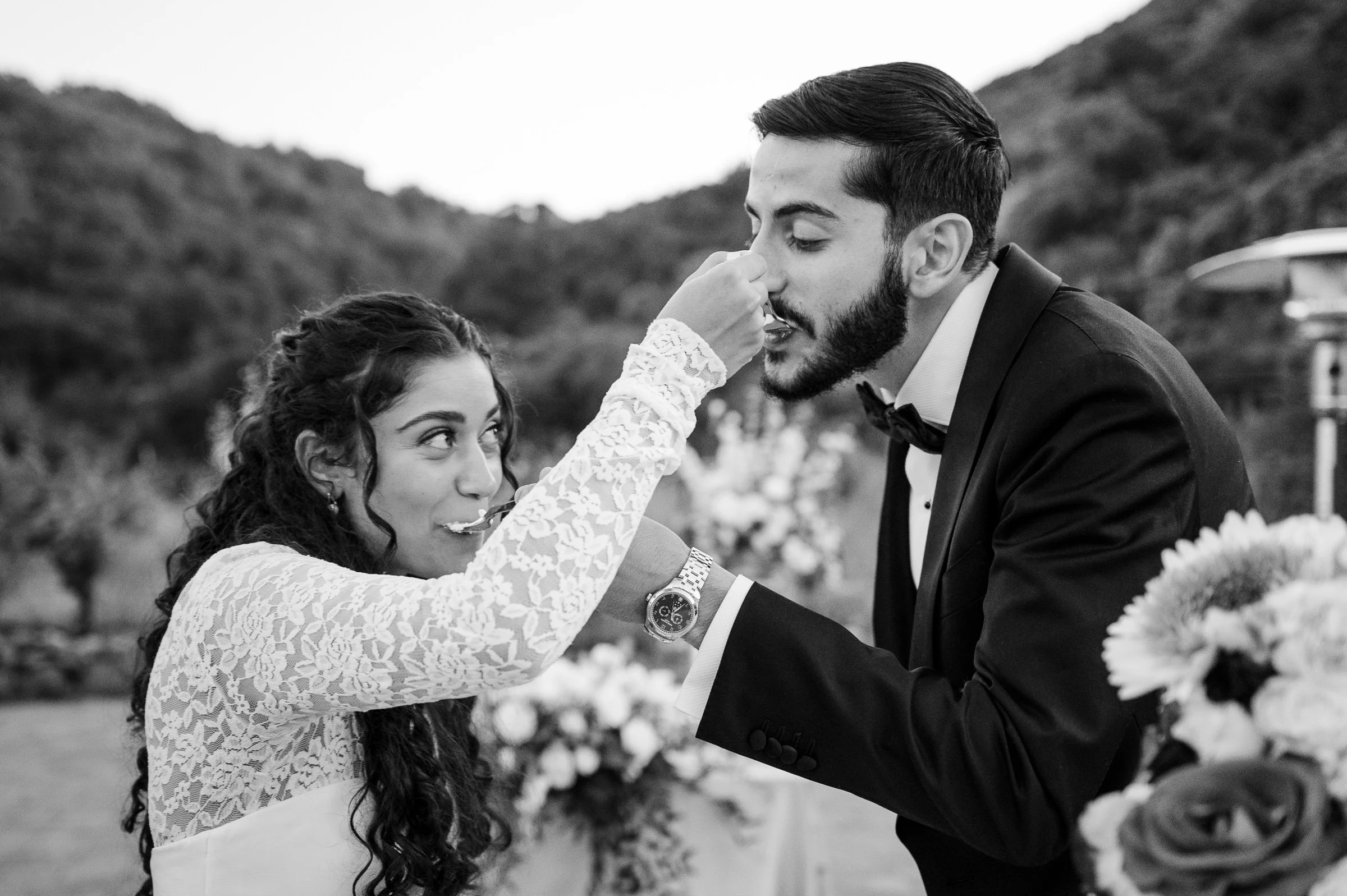 A joyful couple in formal attire feeds each other cake at an outdoor wedding, surrounded by floral arrangements and hills in the background.