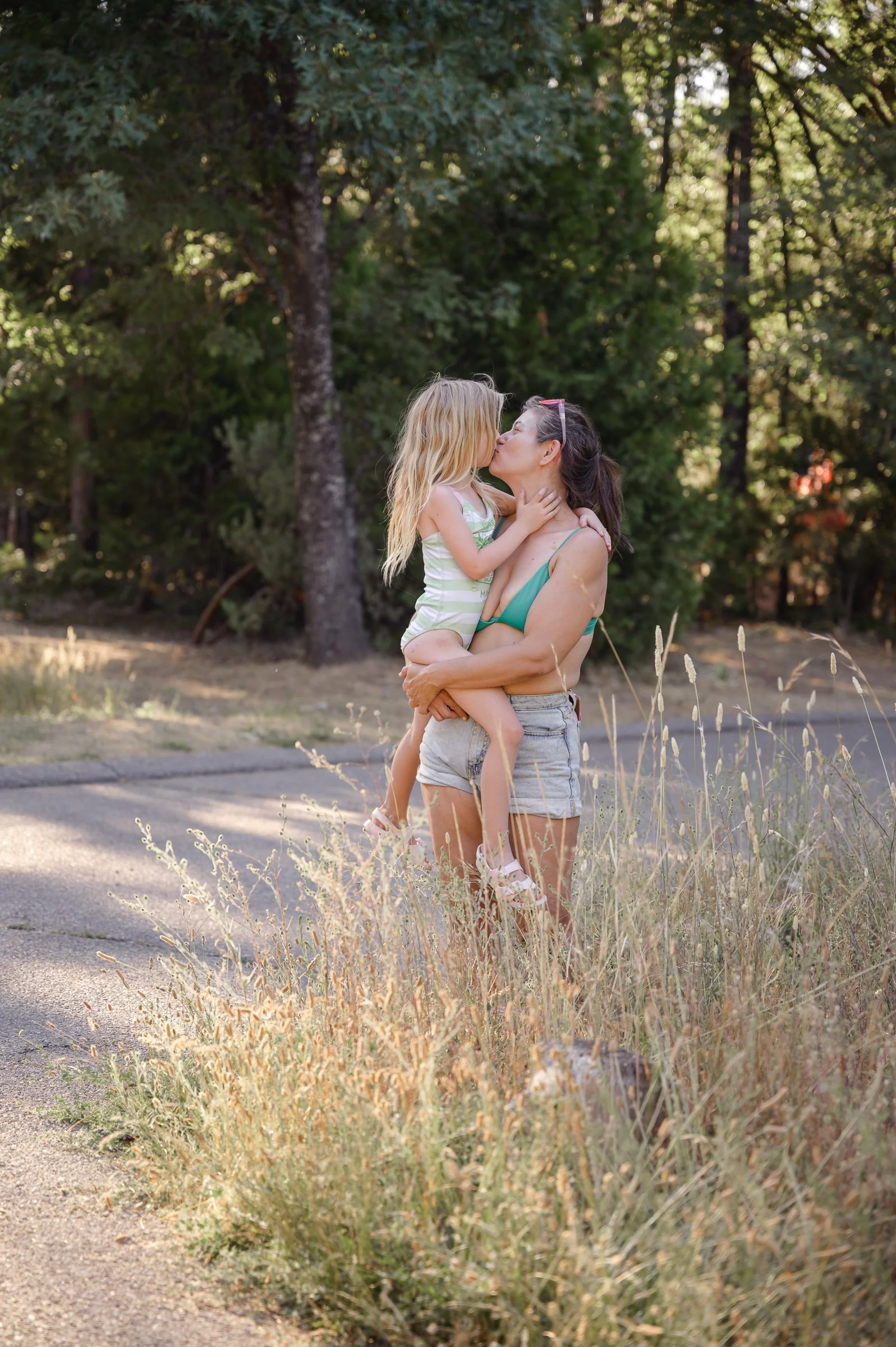 A woman carrying a young girl in her arms outdoors, sharing a kiss, with trees and dry grass in the background.