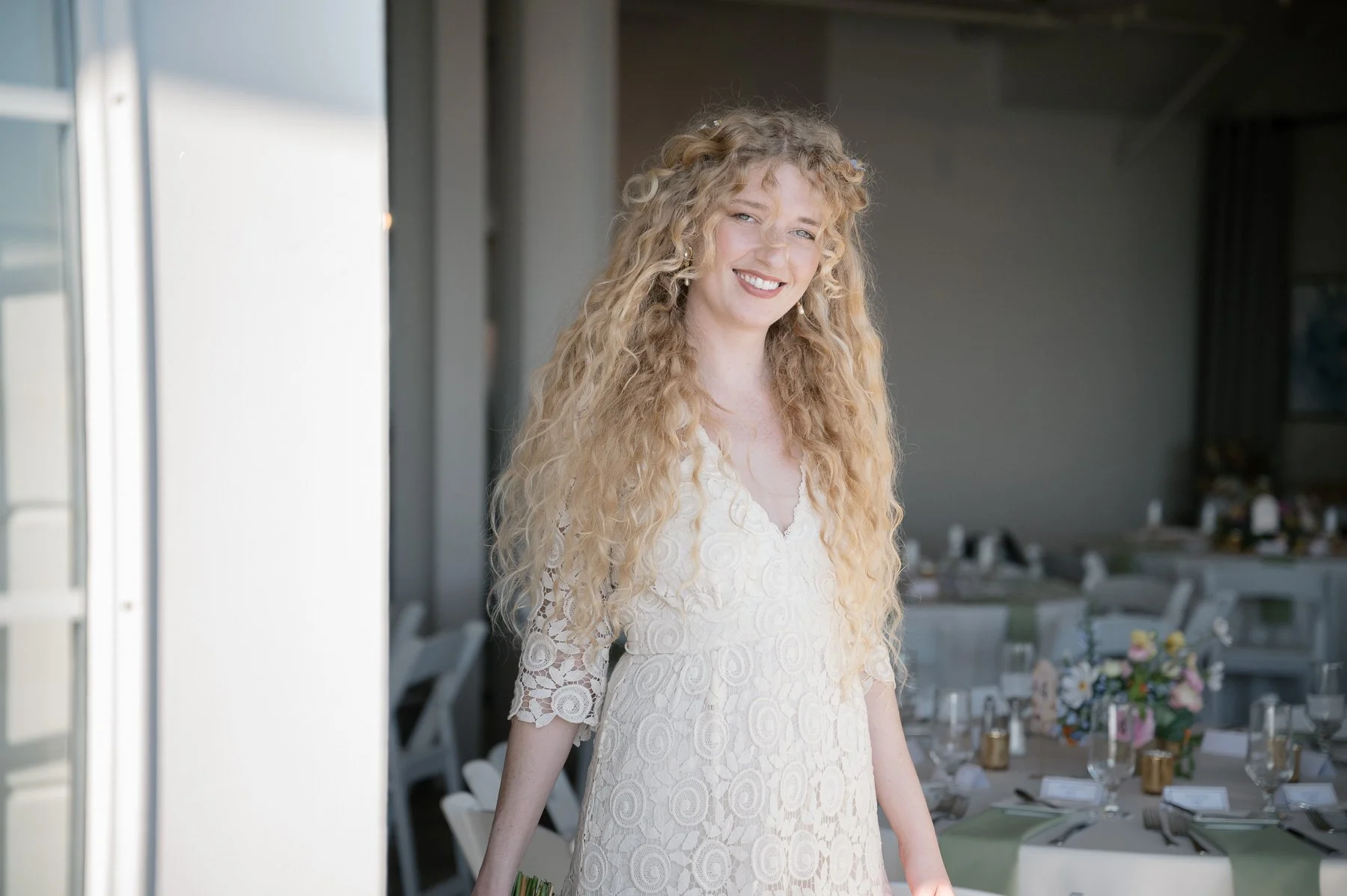 Smiling woman with curly blonde hair in a lace dress in a brightly lit room with elegantly set tables, conveying a joyful and celebratory mood.