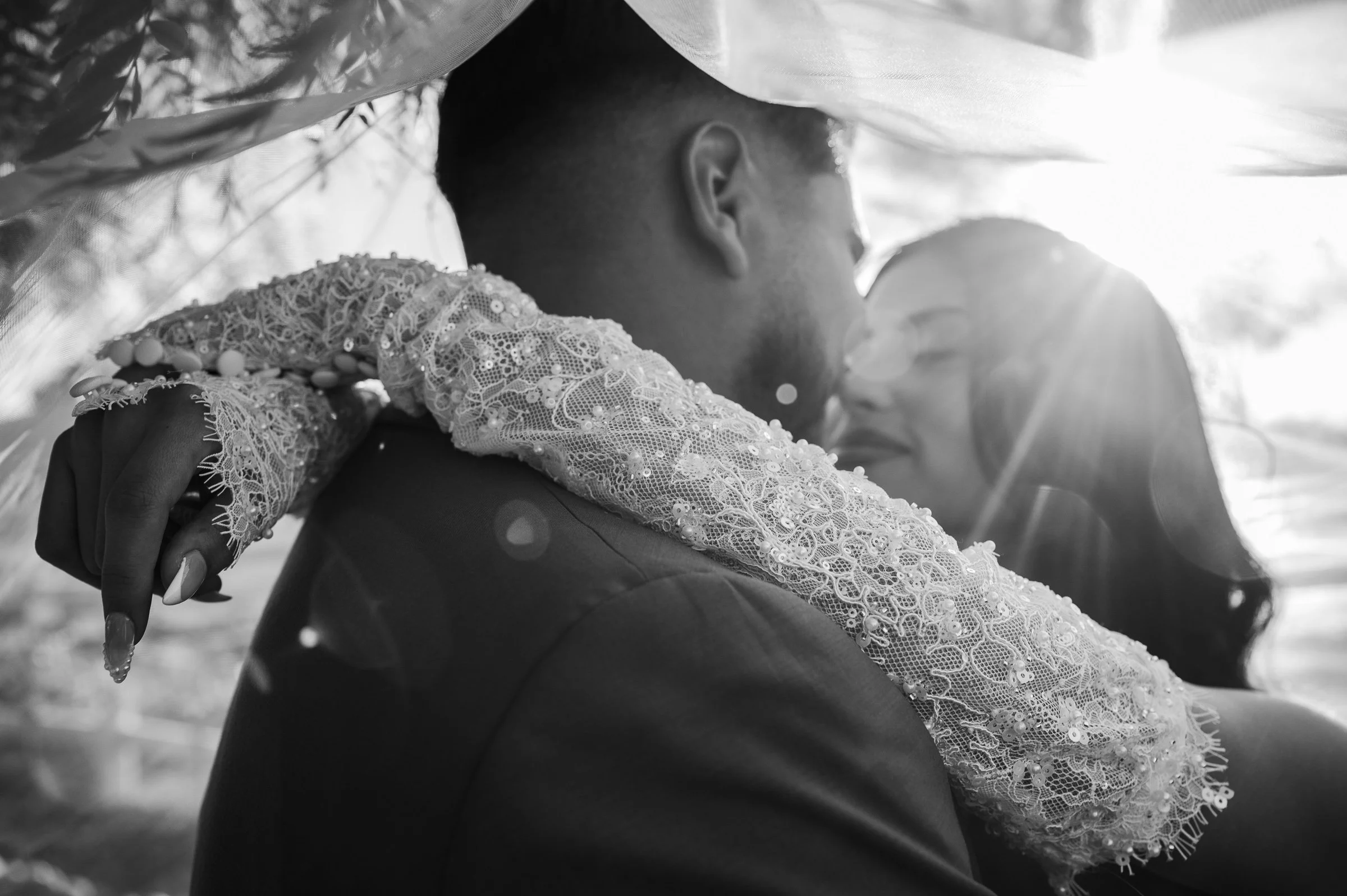 A black-and-white photo of a couple embracing under a veil. The woman's lace-sleeved arm rests on the man's shoulder. Sunlight creates a warm, intimate glow.