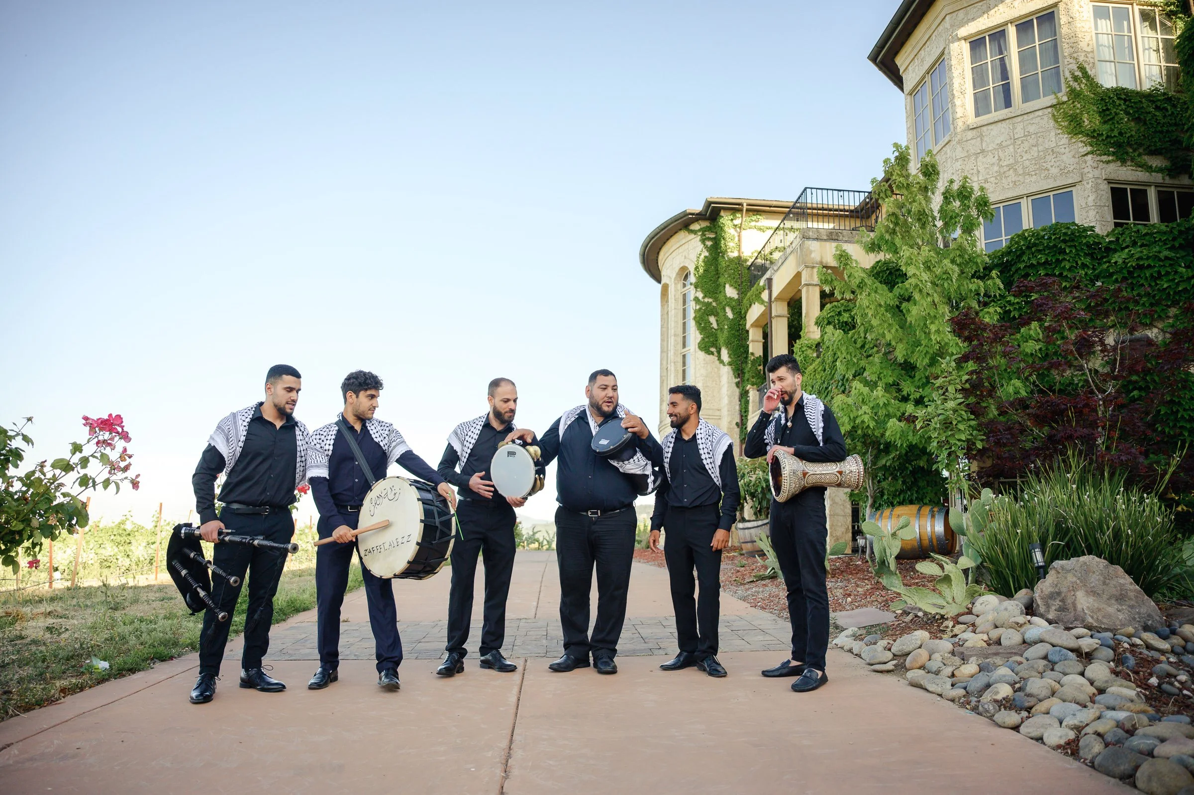 A group of six men in black outfits stand on a path, each holding musical instruments. They're near a building with greenery, under a clear blue sky in preparation for a traditional Palestinian Zaffe.