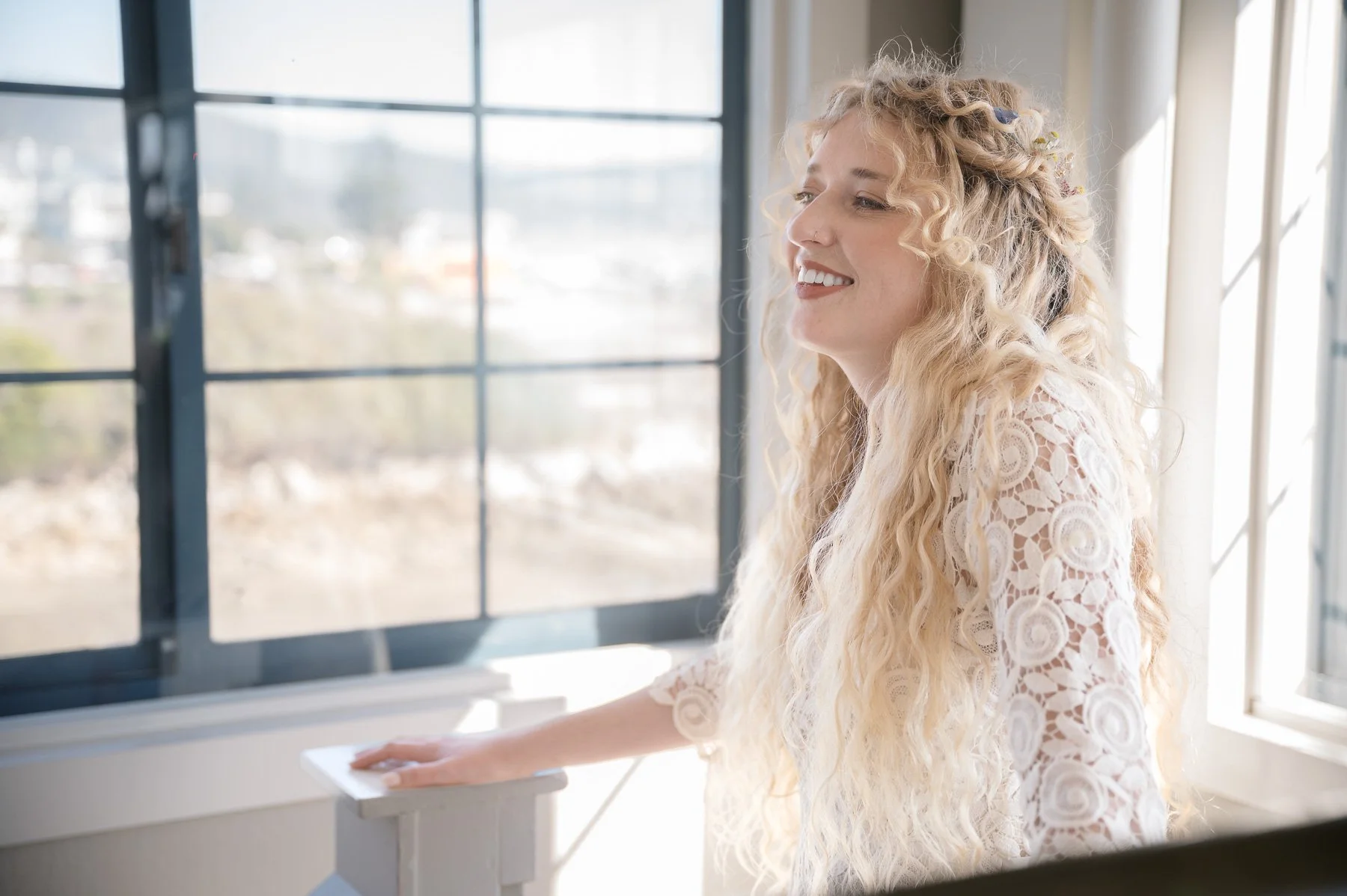 A woman with long, curly blonde hair and a floral lace top smiles while standing by a sunlit window, creating a warm and cheerful atmosphere.