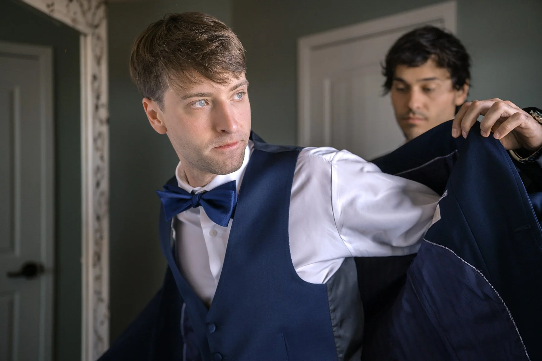 A man in a blue suit and bowtie is being helped into a jacket by another person. He looks focused. The setting is a room with neutral colors.