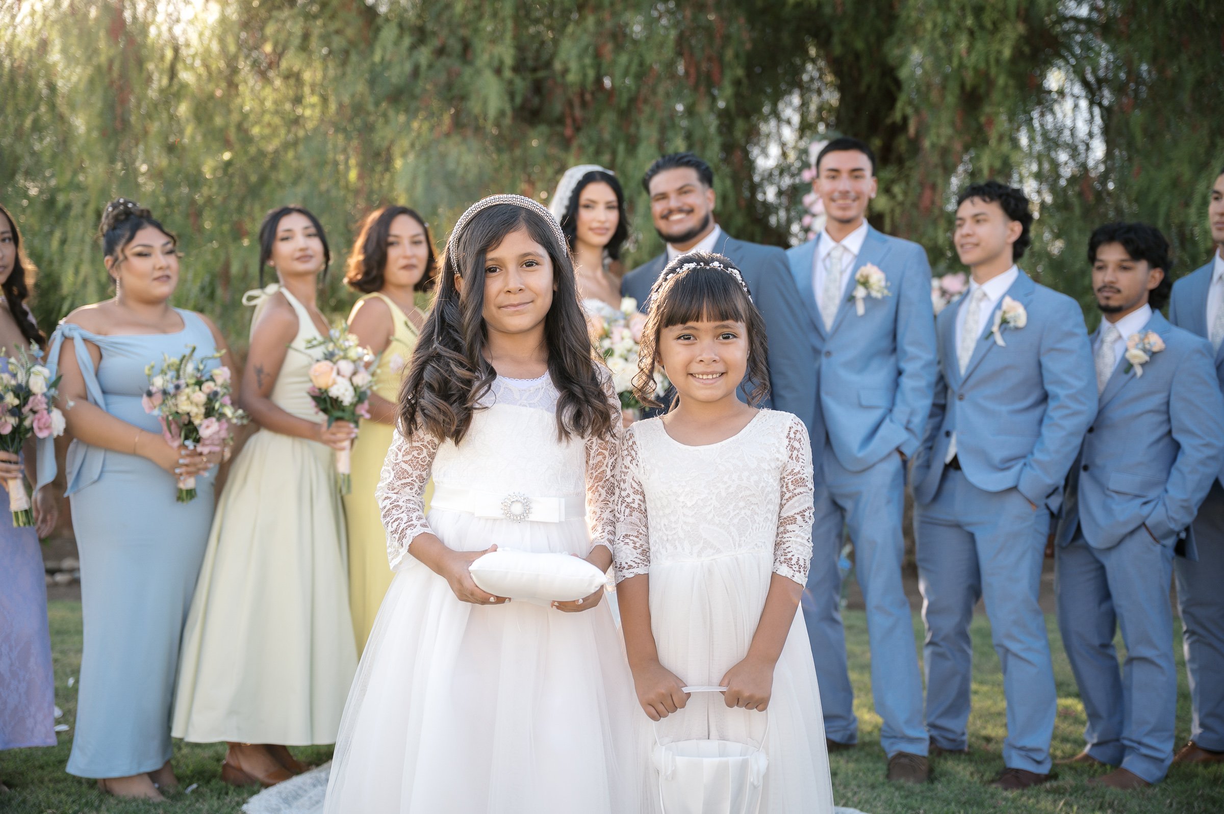Two young flower girls in white dresses stand smiling in front of a wedding party. Bridesmaids in pastel dresses and groomsmen in blue suits are in the background. The setting is outdoors with greenery, creating a joyful and elegant atmosphere.