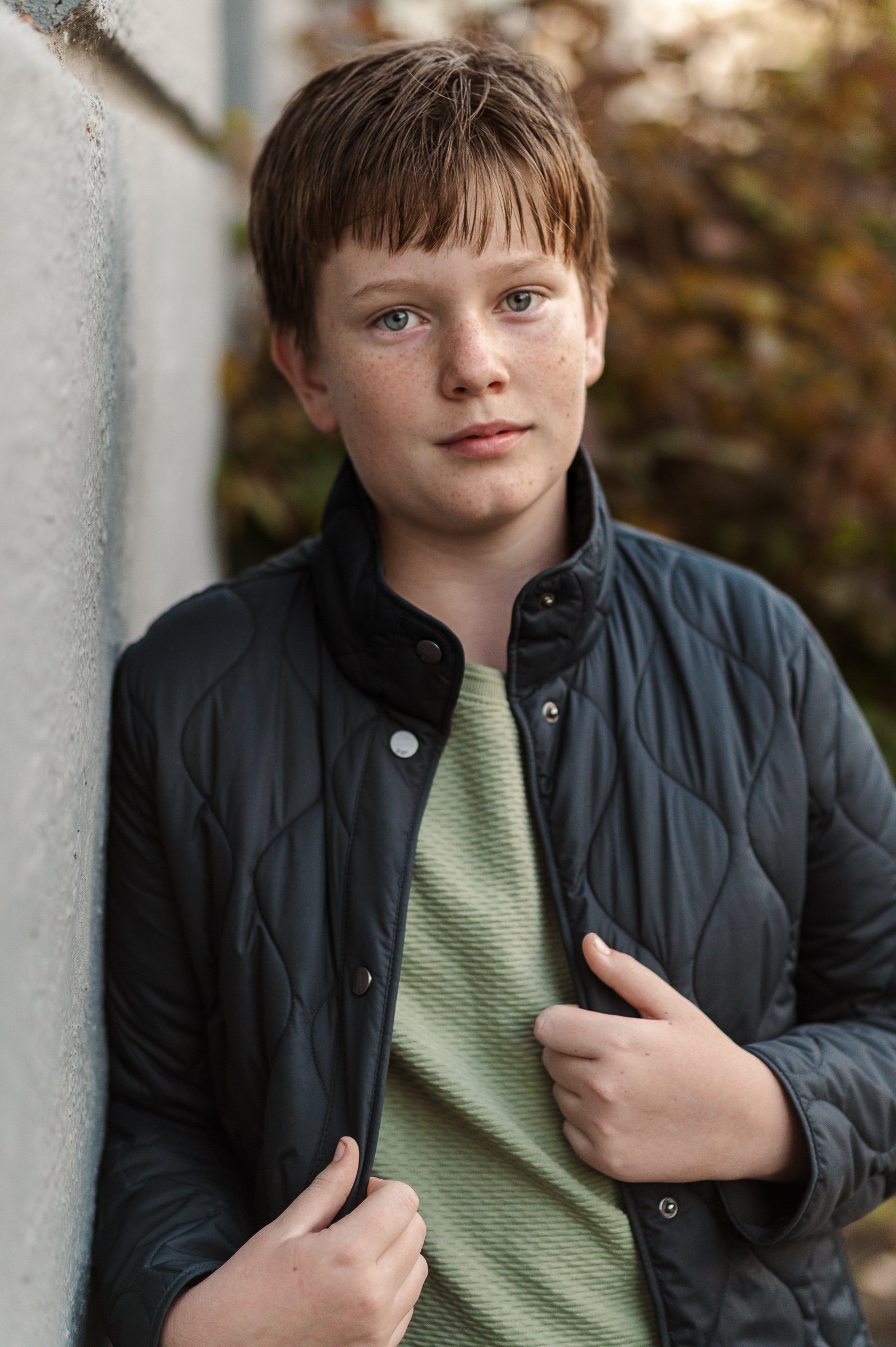 A young boy with red hair and blue eyes, wearing a black quilted jacket and a green striped shirt, stands outdoors near a wall with blurred autumn foliage in the background.