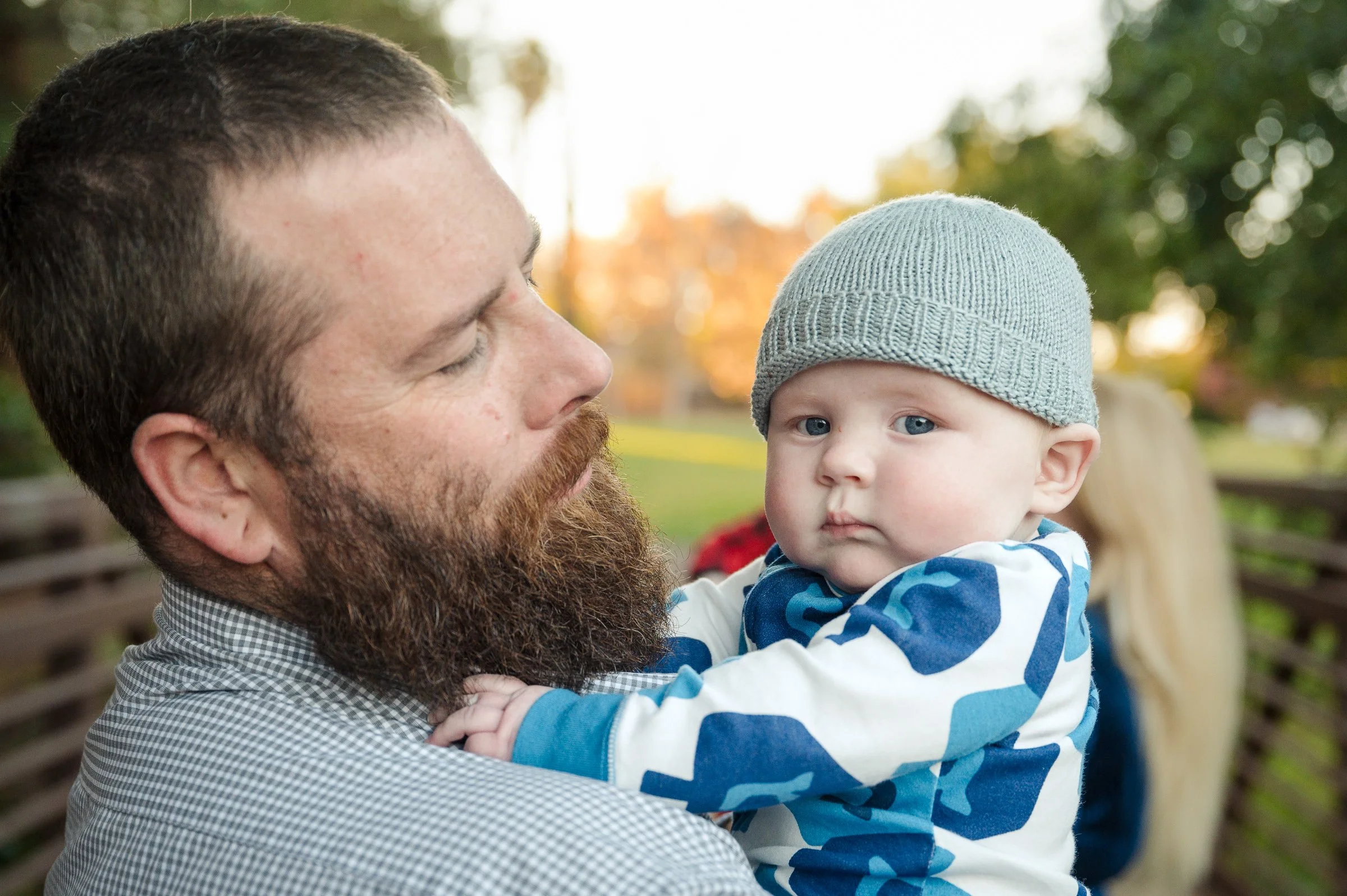 A man with a beard holding a young child with fair skin and blue eyes, wearing a gray knit hat and a blue and white striped shirt, outdoors with blurred trees and sunset in the background.