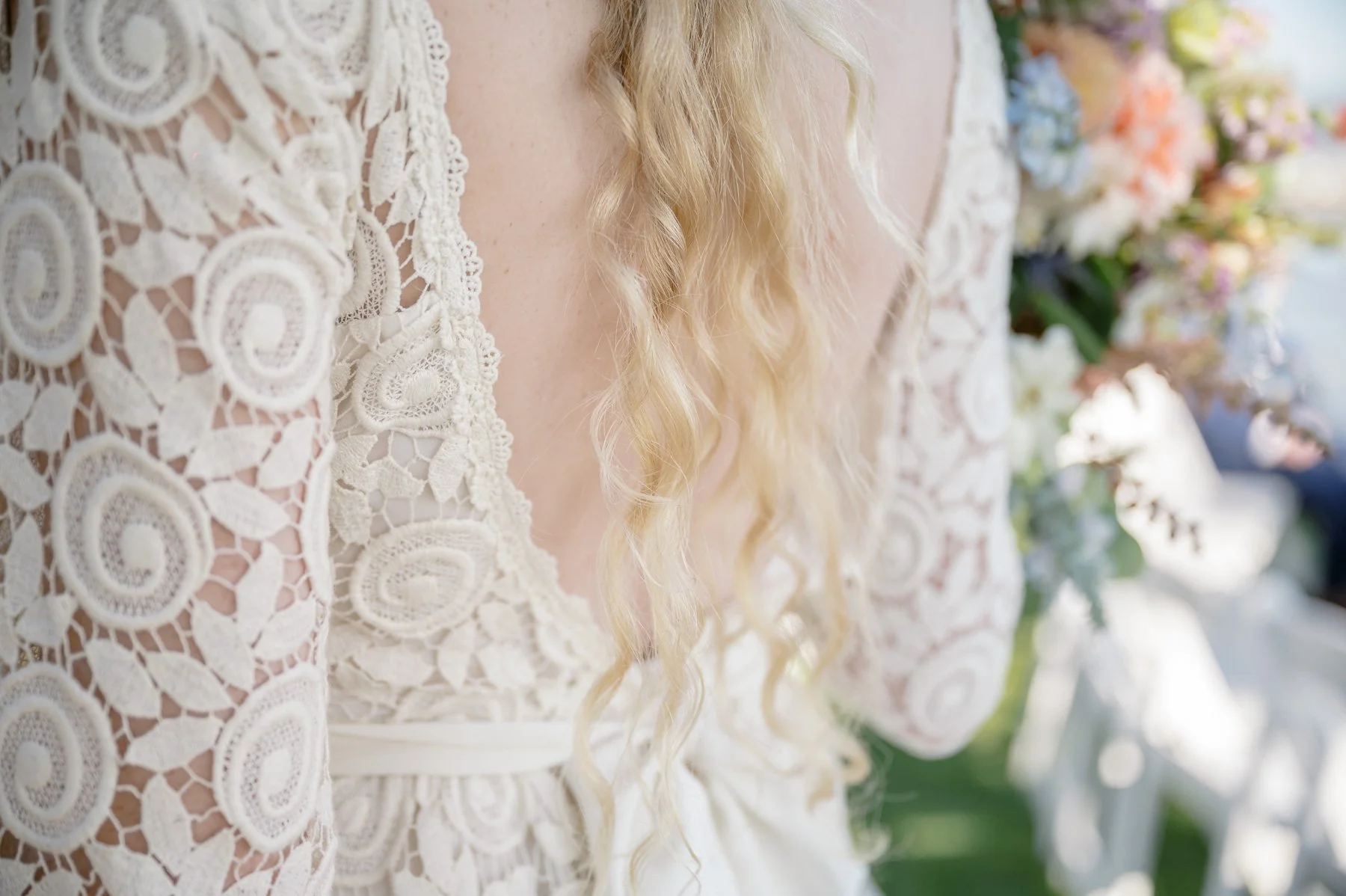 Close-up of a woman in a lace wedding dress with intricate circular patterns and wavy blond hair. She holds a colorful bouquet, conveying a romantic atmosphere.