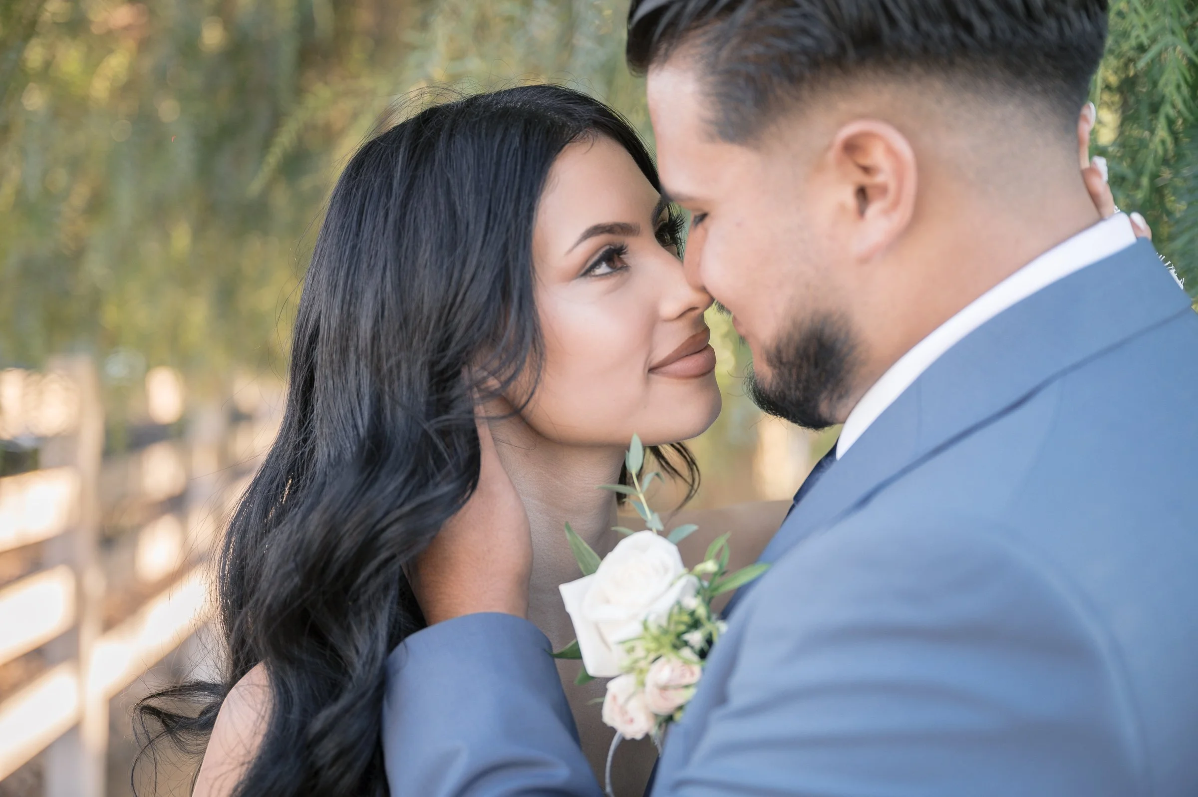 A couple shares an intimate moment, gazing into each other's eyes. The man, in a blue suit, holds her face; she wears a white rose corsage. Romantic and serene.