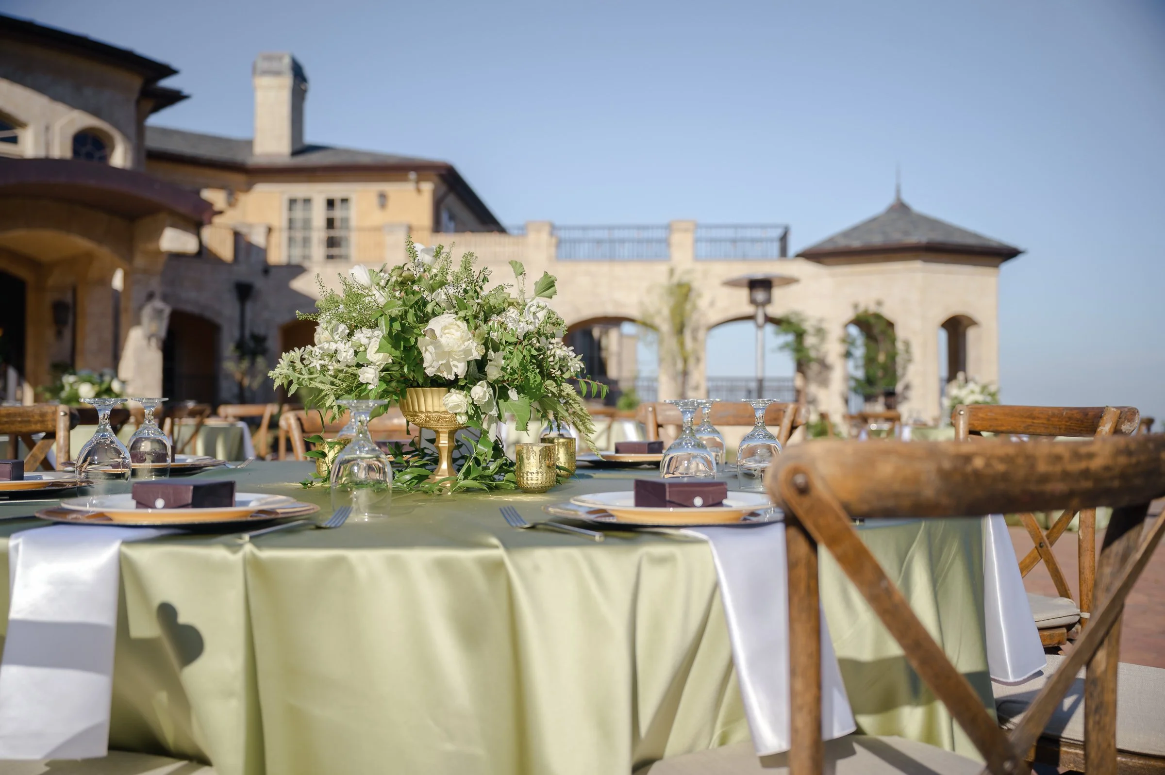 Elegant outdoor table setting with green tablecloth, wooden chairs, and floral centerpiece. Stone villa in background under clear blue sky.