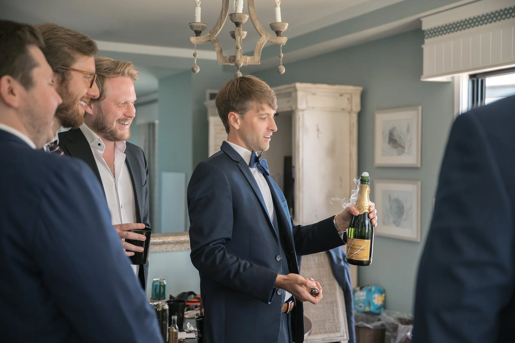 A group of men in suits are gathered indoors, smiling as one holds a champagne bottle. The scene conveys celebration and camaraderie.