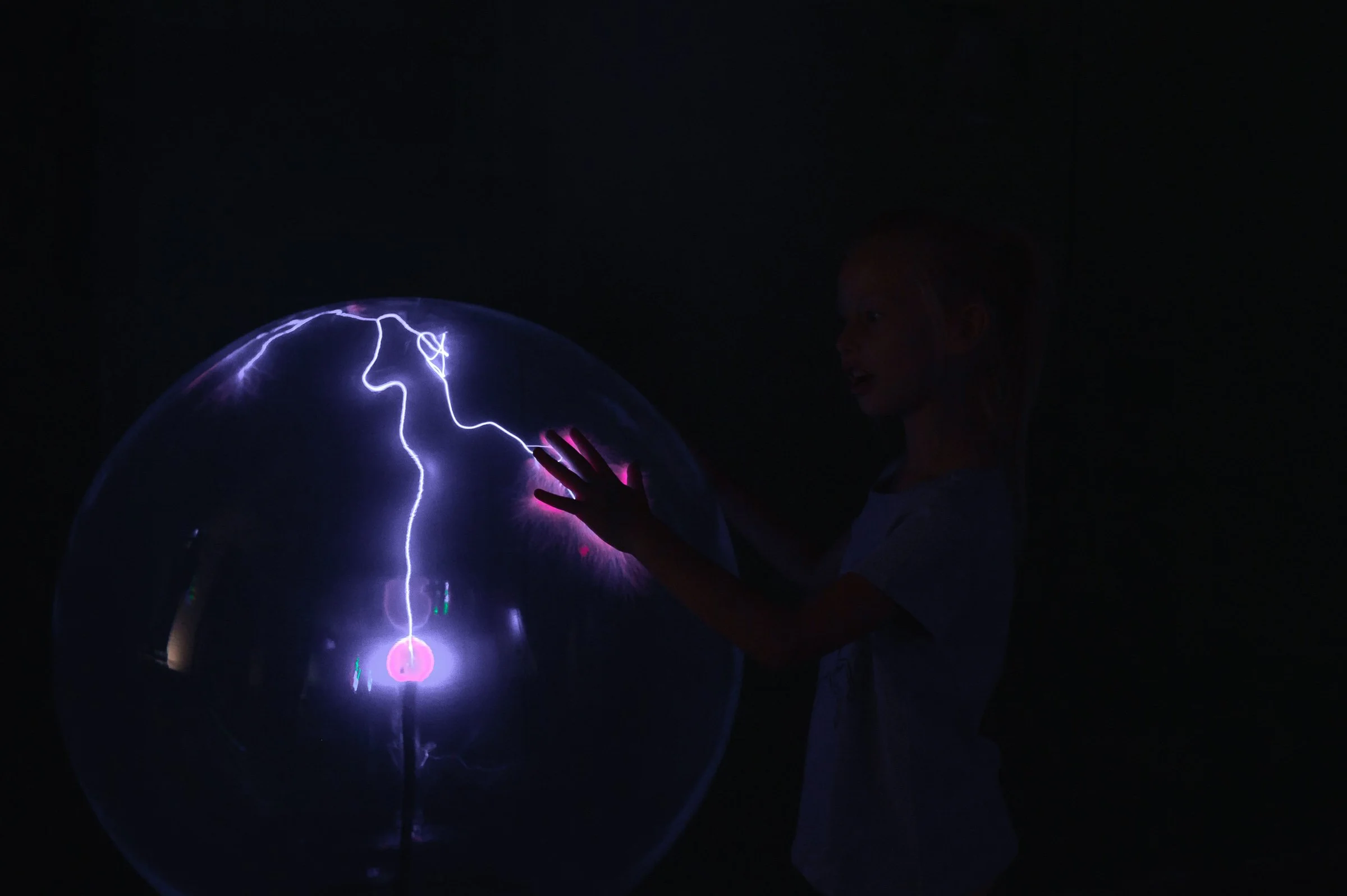 A young girl smiling and touching a spherical Tesla coil that produces visible lightning discharges in a dark environment.