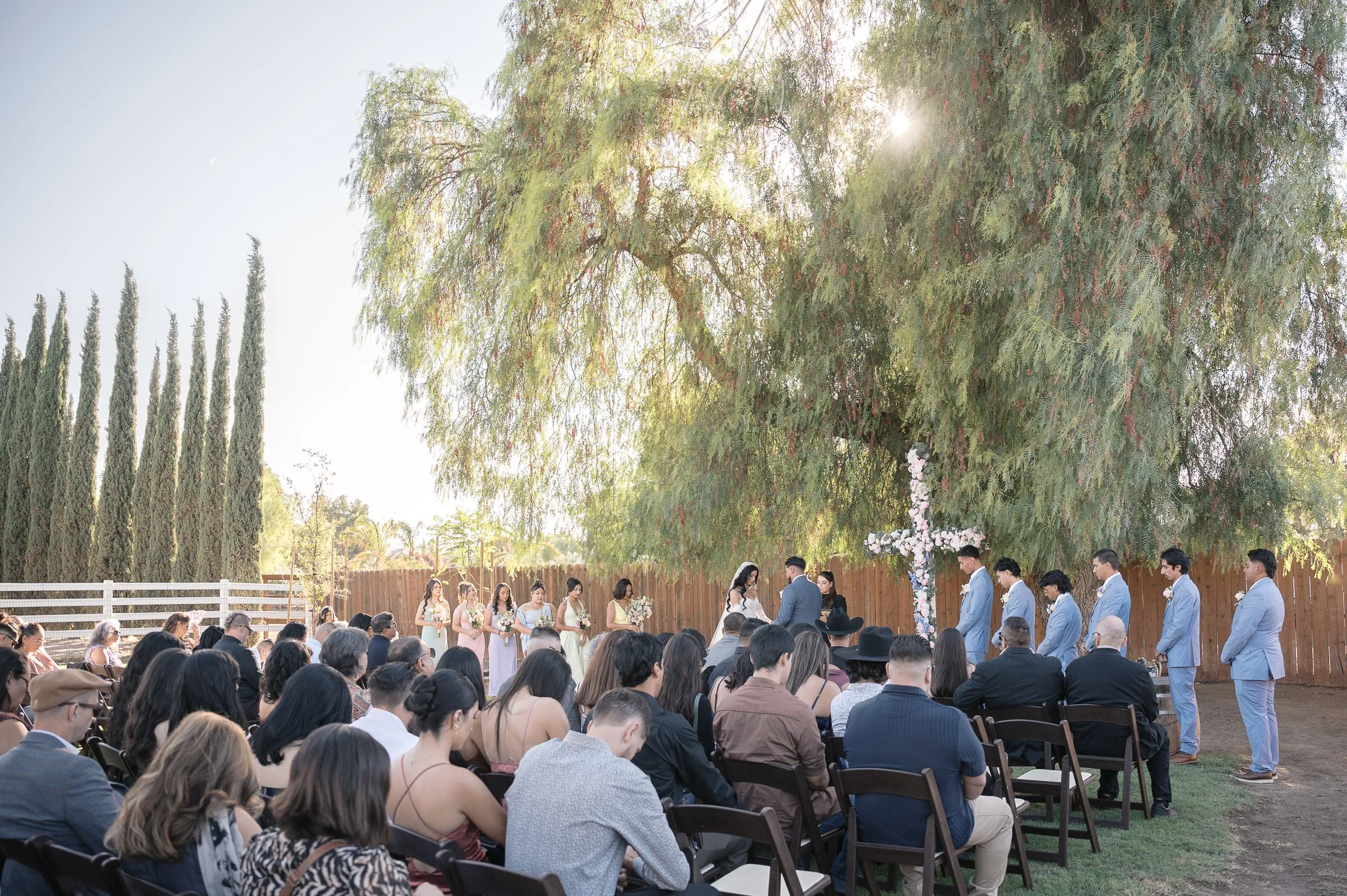 Outdoor wedding ceremony under a large tree with sunlight filtering through. The bride and groom stand by a flowered cross, surrounded by guests and attendants.