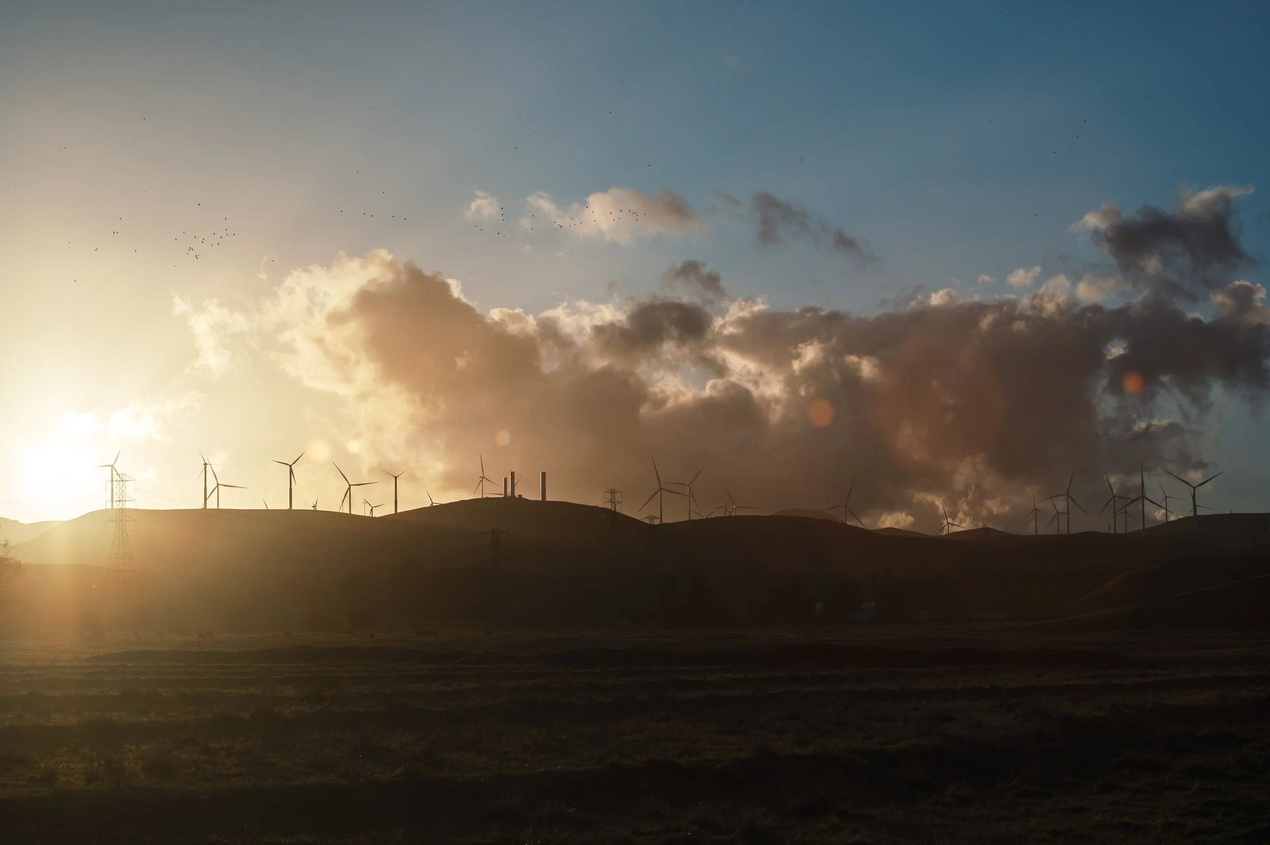 A landscape view at sunset with wind turbines on rolling hills and a partly cloudy sky.