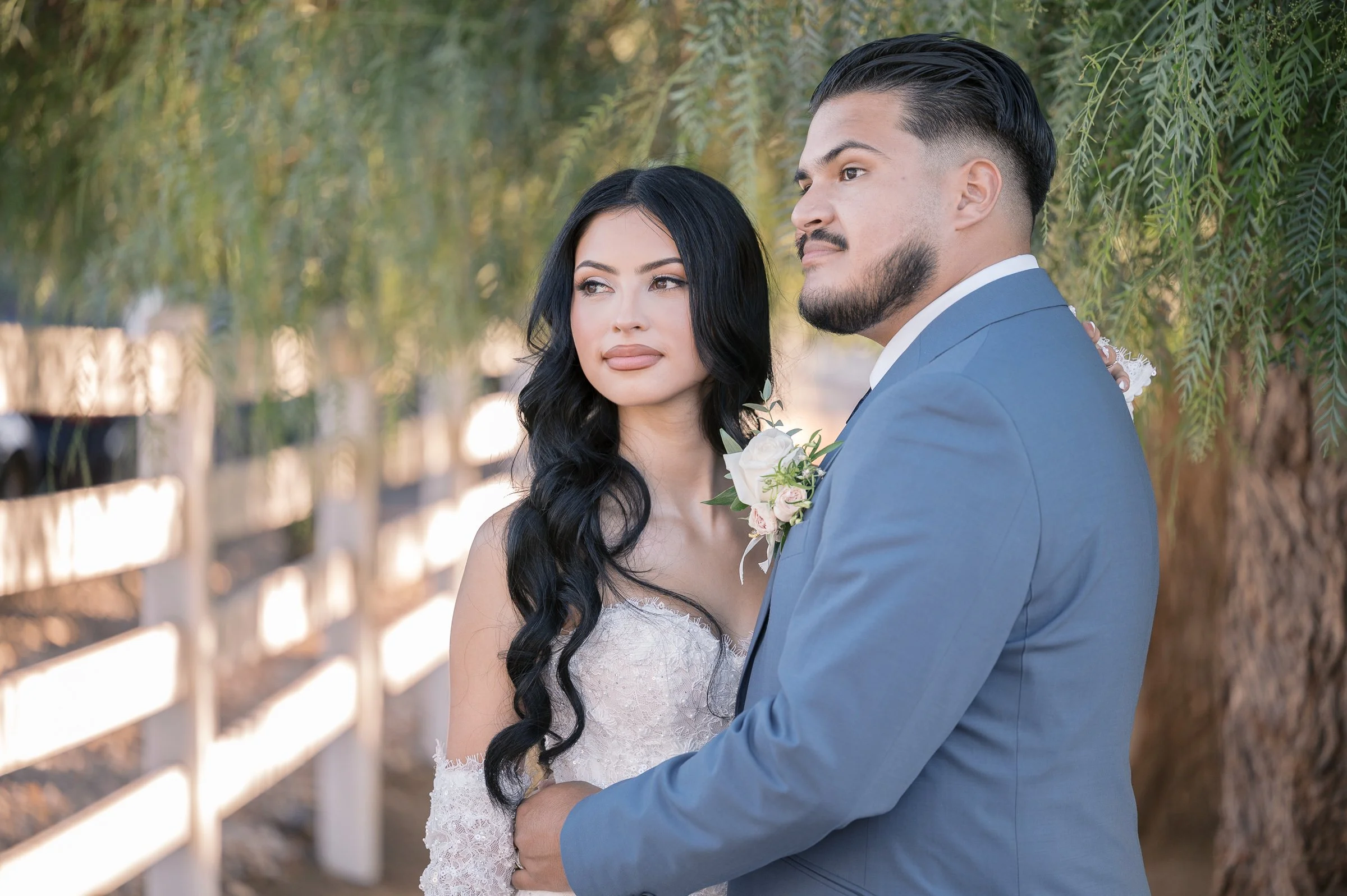 A couple stands together outdoors in soft lighting. The woman wears a lacy dress, and the man wears a blue suit with a boutonniere. They appear serene and content.