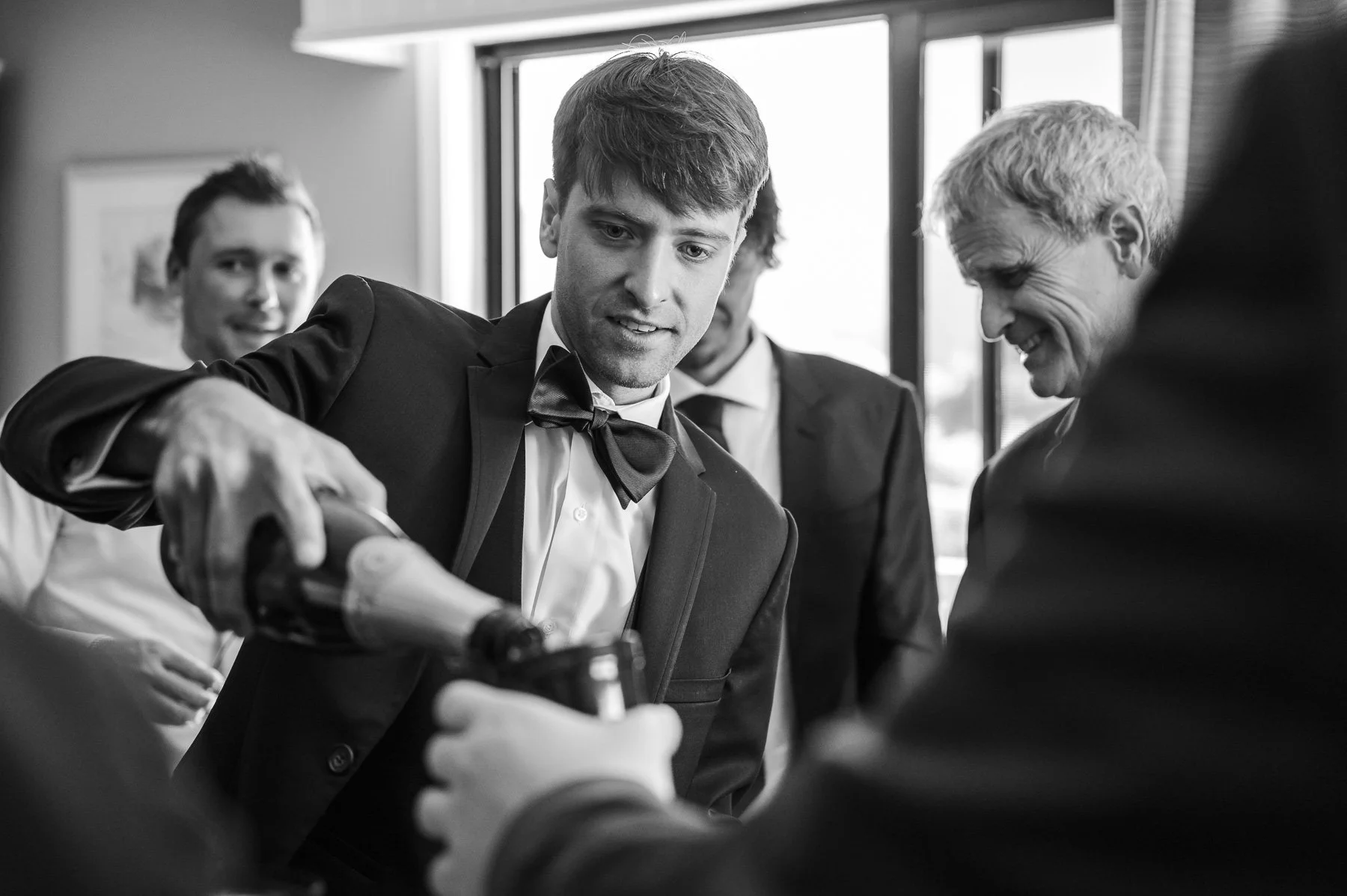 A man in a suit and bow tie pours champagne into a glass held by another person, surrounded by smiling men in suits, creating a celebratory atmosphere.