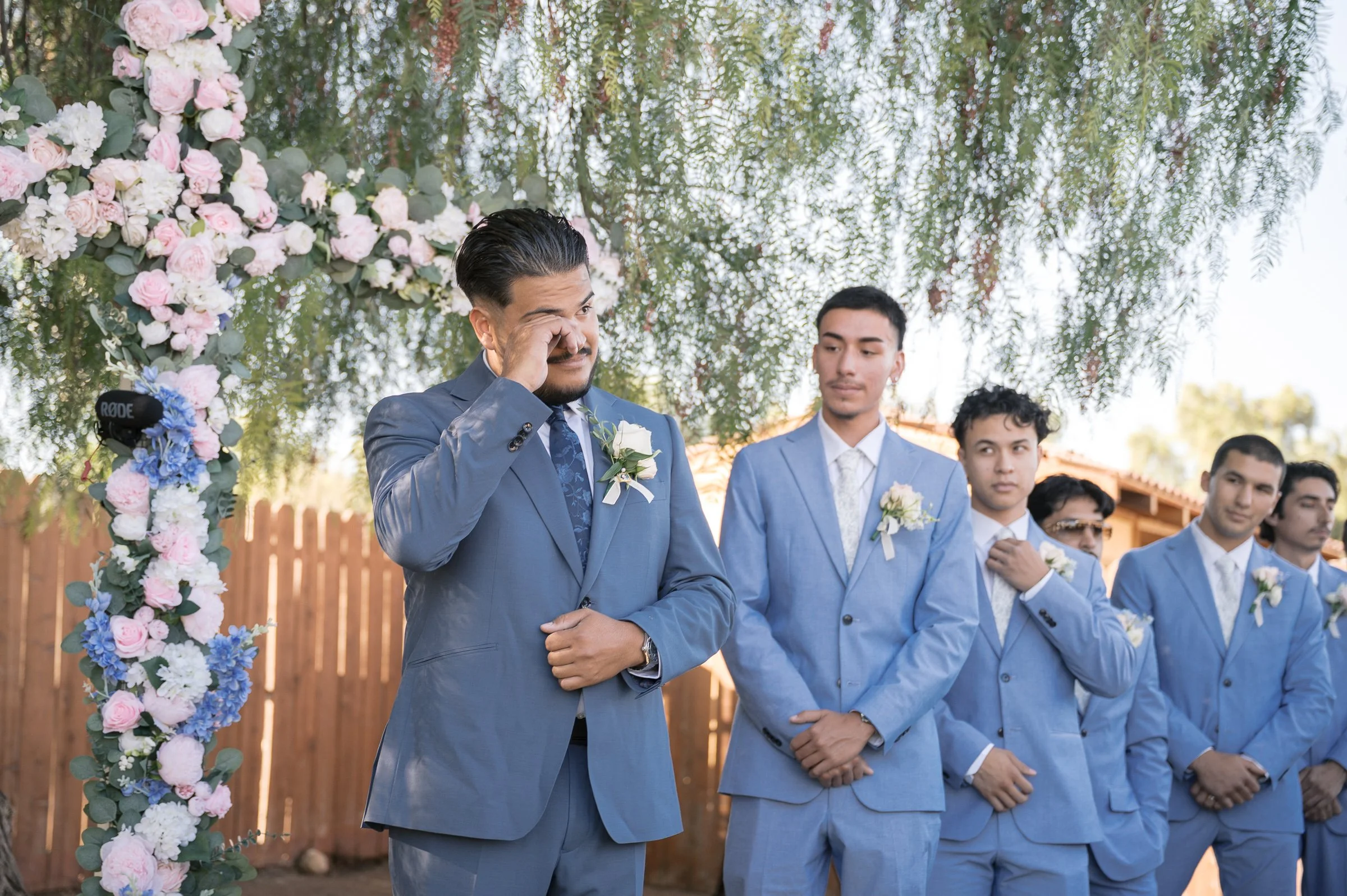A groom in a blue suit wipes a tear beside a floral cross, attended by groomsmen in matching suits. The scene is emotional and heartfelt.