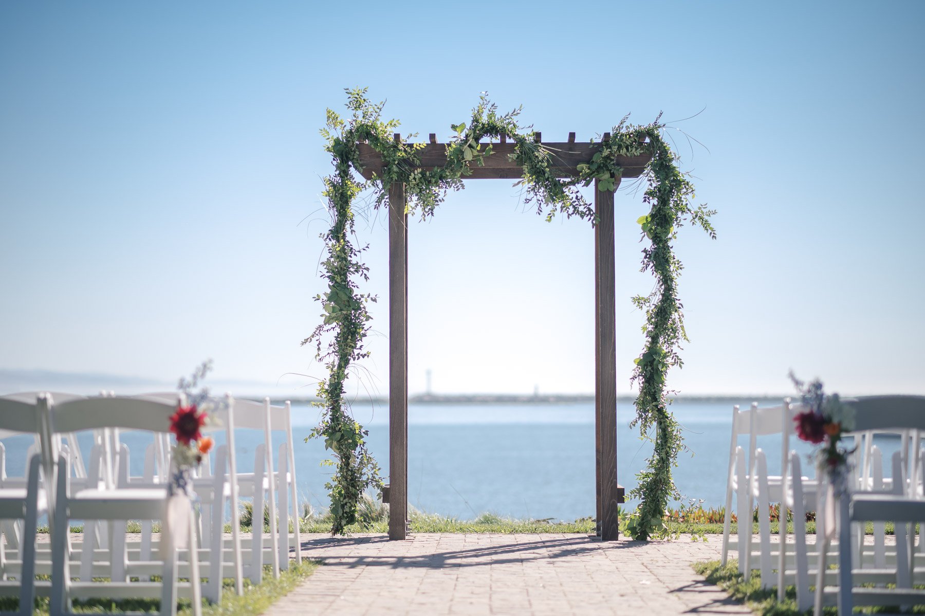 Wooden wedding arch adorned with greenery, overlooking a calm blue sea. White chairs with floral accents line both sides under a clear sky.