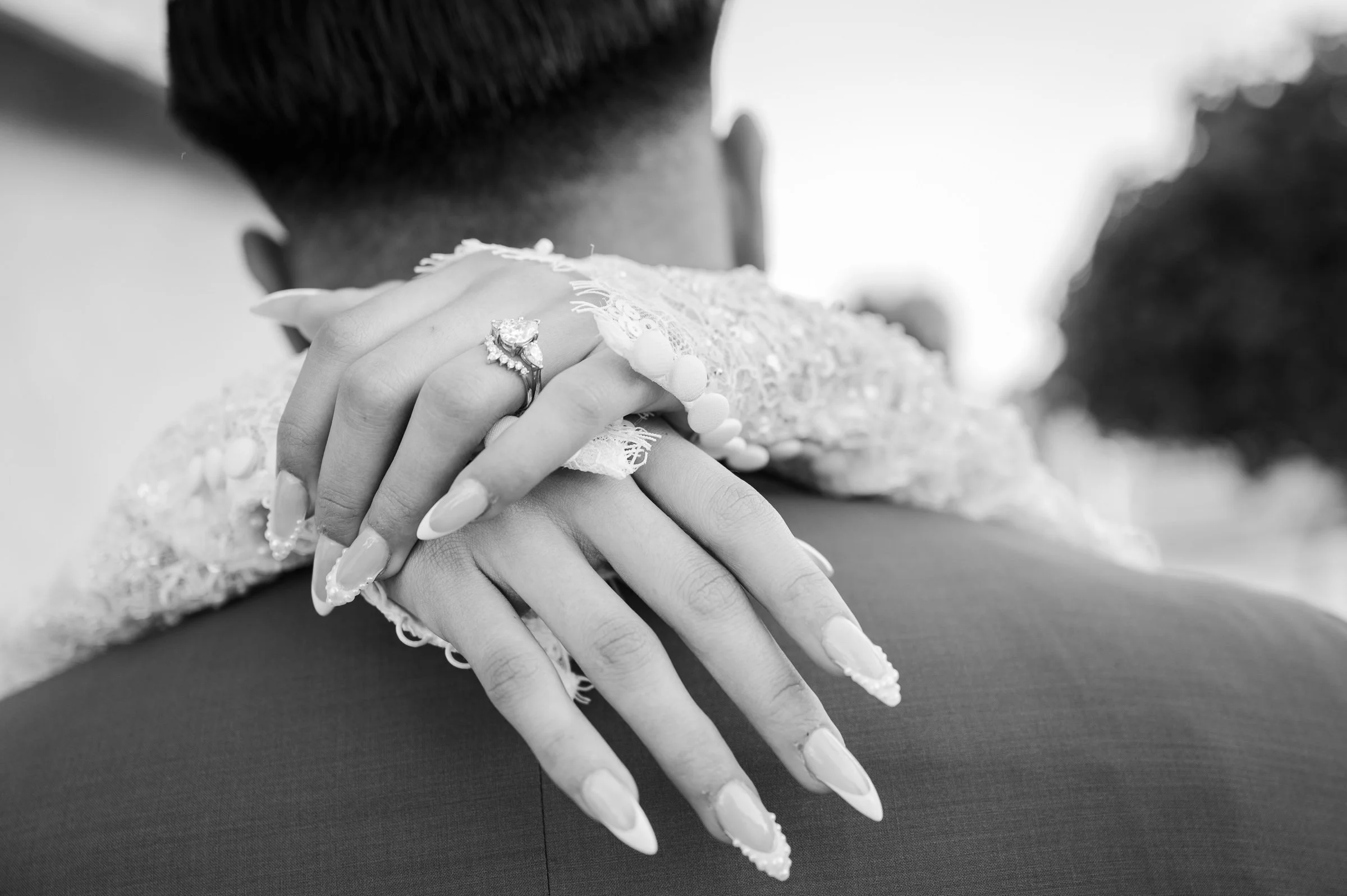 A close-up in black and white showing a bride's hands with an ornate ring resting on a groom's shoulder, creating an intimate and loving atmosphere.