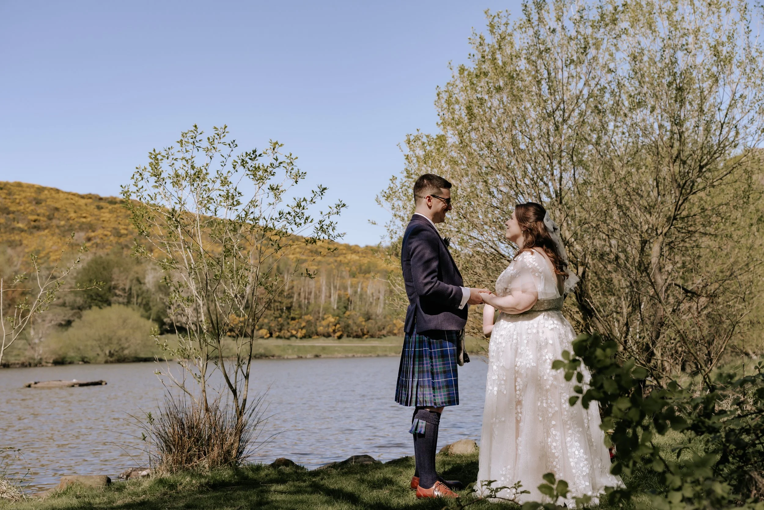 A bride and groom holding hands and smiling at each other outdoors by a loch, surrounded by trees and hills in the background.
