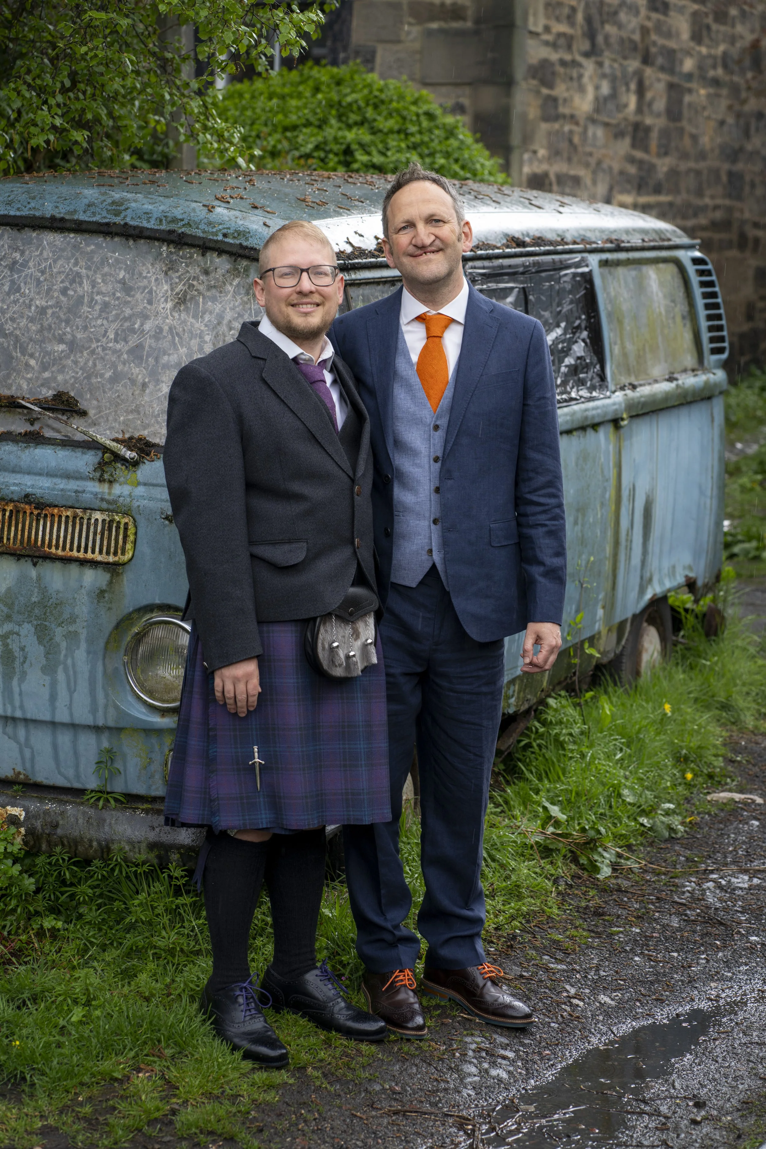 A male couple dressed in wedding attire standing outdoors on a rainy day. They are standing next to an old, rusty blue vehicle with broken windows and overgrown grass around it.