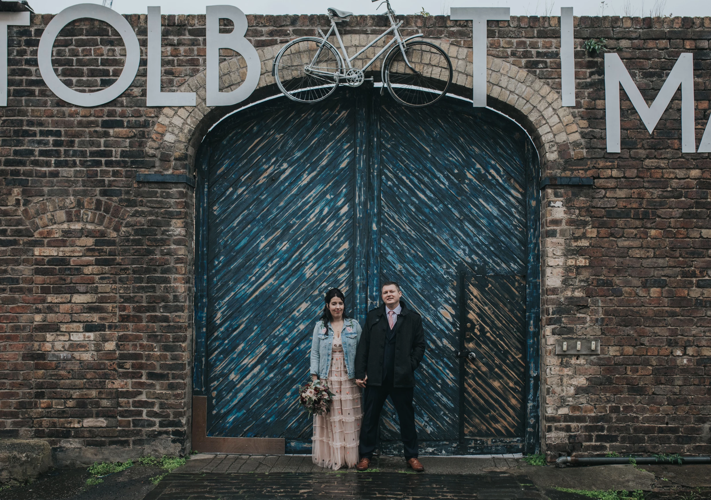 A bride and groom holding hands and standing in front of a large painted door with brick walls, with a bicycle sculpture and some lettering on the wall above.