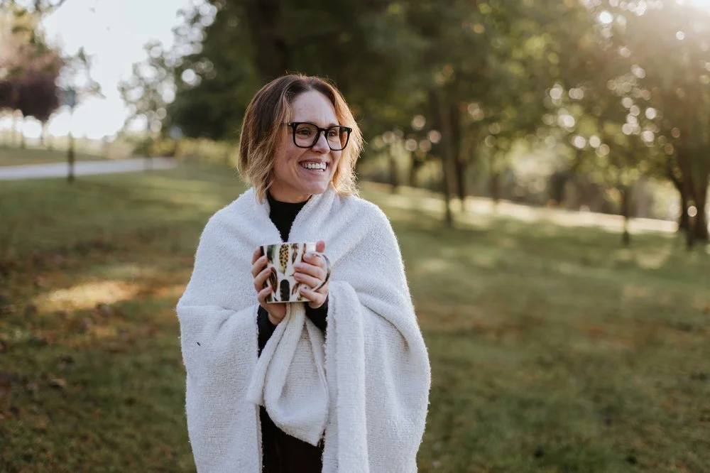 Amber Harris wrapped in a cozy white blanket, holding a mug and smiling warmly in a sunlit park